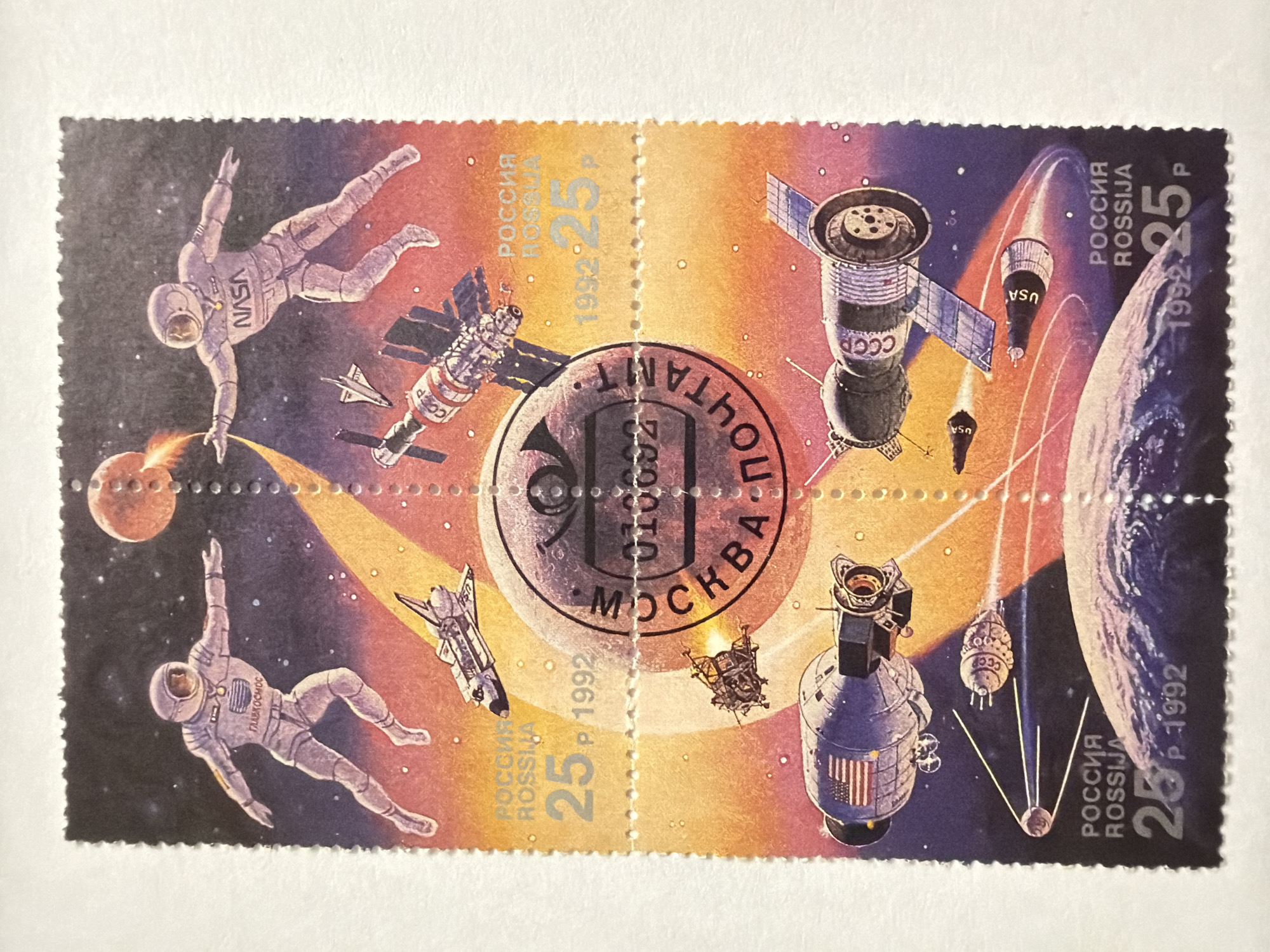 The image is a block of four Russian postage stamps issued in 1992 depicting various aspects of space exploration. The stamps are connected and feature a central postmark stamp.
The individual stamps contain the following imagery:
* **Top Left:** Two astronauts floating in space with a planet and a spacecraft. One astronaut's spacesuit has "NASA" on it, while the other's says "ГЛАВКОСМОС" (Glavkosmos).
* **Top Right:** A spacecraft, along with a vehicle marked "USA" and the Earth in the background.
* **Bottom Left:** Similar to the top left, another astronaut floats in space with a spacecraft.
* **Bottom Right:** A different spacecraft with the American flag on it, a vehicle marked "CCCP," and the Earth with a vehicle marked "USA."
All four stamps have "РОССИЯ ROSSIJA" and "25 P 1992" printed on them. "28 P 1992" is printed on the bottom right stamp. The location "ОСКВА" (Moscow) is visible within the central postmark.
The description does not include any information regarding Izhevsk, Russia.