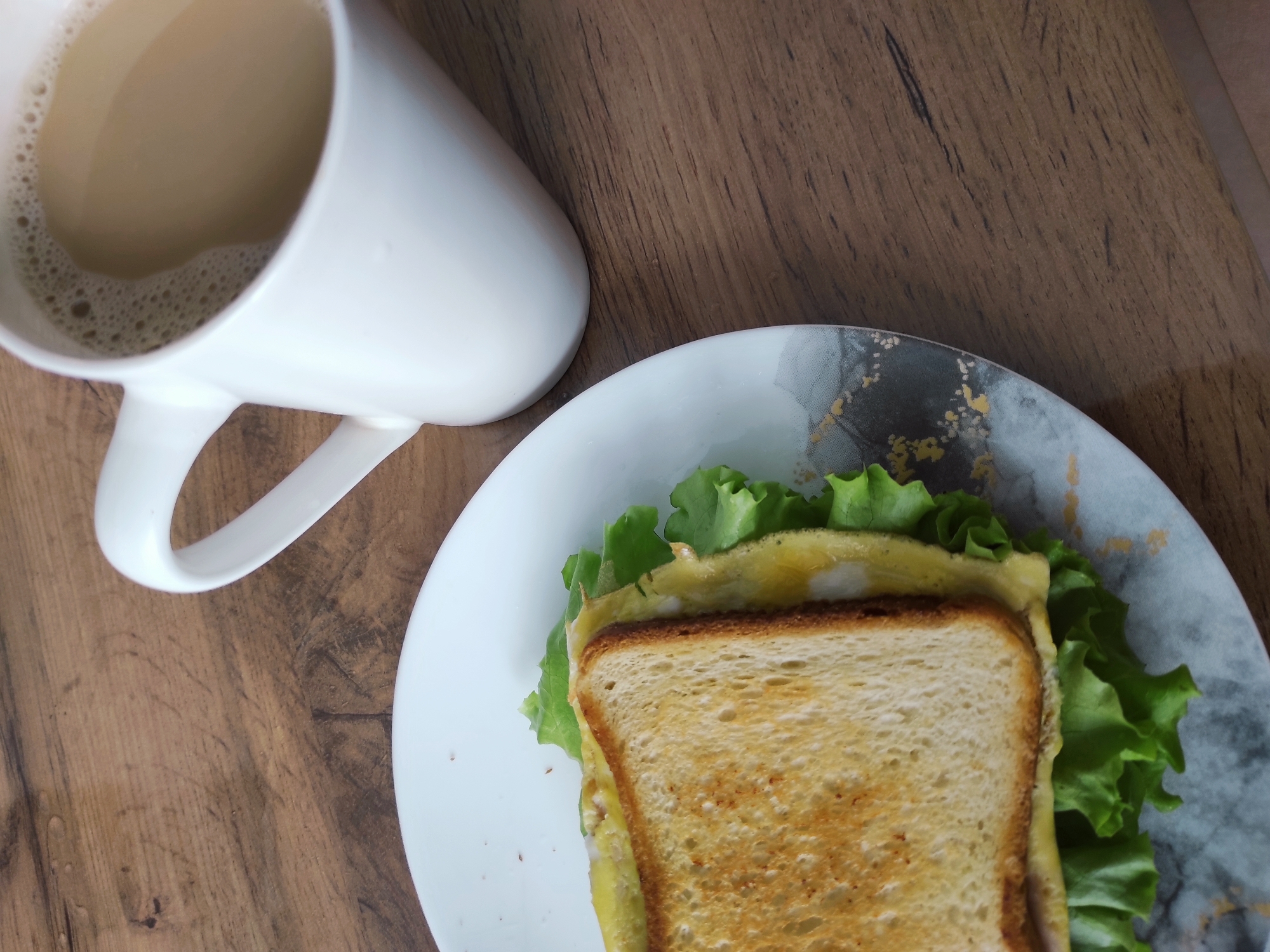 A top-down, slightly angled shot captures a simple breakfast or snack arranged on a light brown wooden table. In the upper left quadrant, a white ceramic mug holds a light brown, foamy beverage, likely coffee or tea with milk. The surface of the drink shows small bubbles. To the bottom right, a white ceramic plate with a distinctive grey and black design, accented with irregular gold flecks, holds a sandwich. The sandwich consists of two slices of lightly toasted bread, a layer of fluffy yellow scrambled egg or omelette, and several fresh green lettuce leaves peeking out from beneath the bread. The wooden table surface features visible grain patterns. The scene suggests an indoor setting, possibly a home in Chaikovsky, Russia. No people are visible, and there is no discernible text.