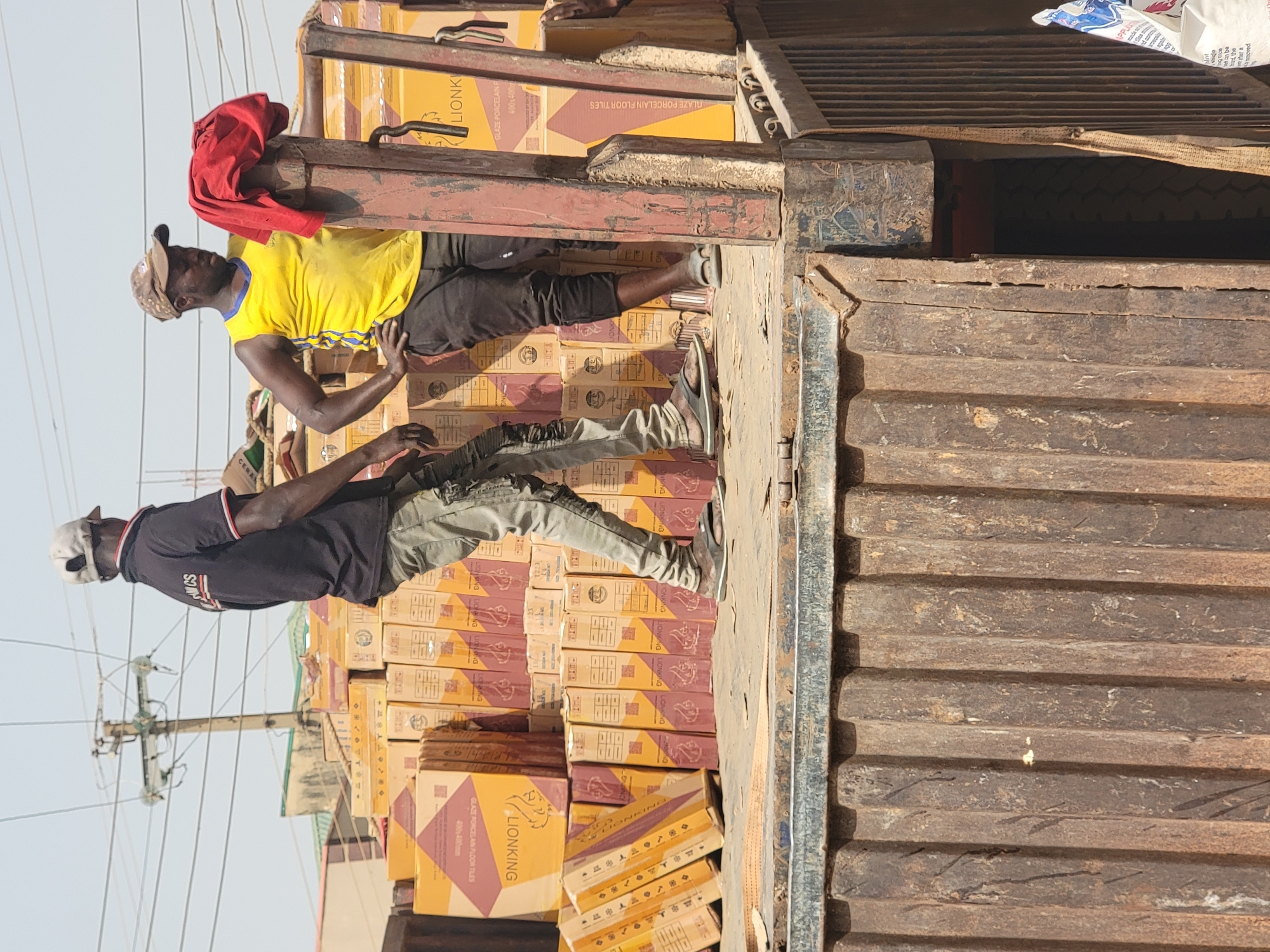 The image shows two men on top of a truck loaded with boxes of Lionking brand floor tiles, likely in Maiburiji, Nigeria. The truck bed is metal with ribbed siding. The man closest to the truck cabin wears a yellow t-shirt, black pants, and a tan baseball cap with a red rag draped on a beam. The other man wears a black t-shirt, gray pants, a white cap, and sandals. The boxes are labeled "LIONKING GLAZE PORCELAIN FLOOR TILES". A utility pole and building are visible in the background.