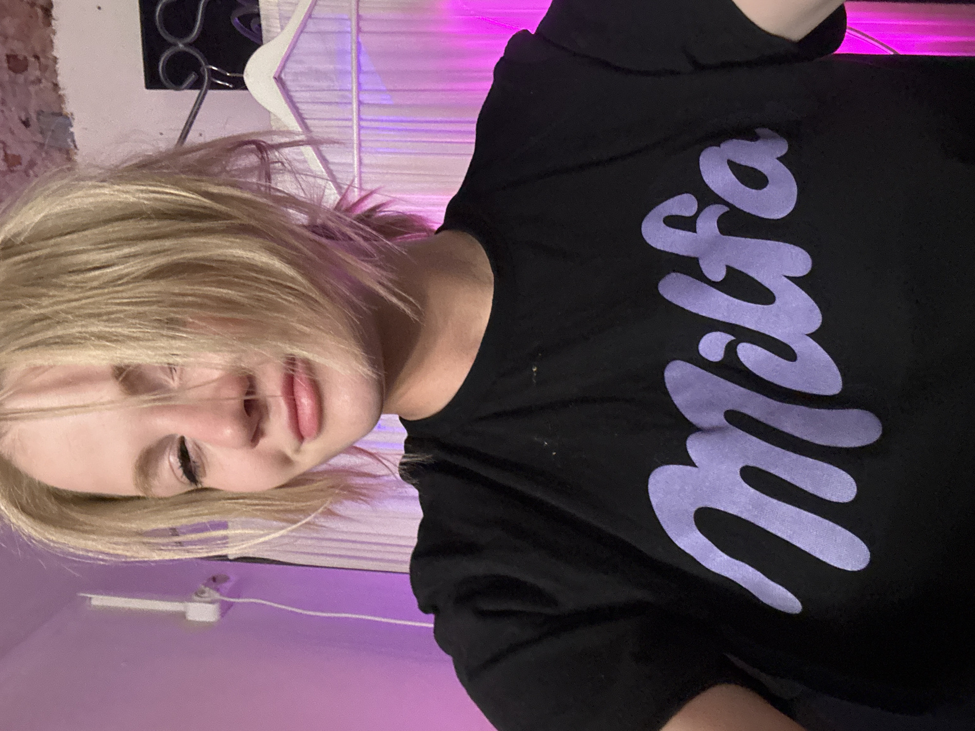 A young woman with blonde hair is wearing a black t-shirt with the word "Milka" printed in purple lettering, angled to the right. The scene has a pink and purple hue, possibly from neon lighting. A wire hanger is visible behind her, along with a blurred brick wall in the background. An electrical outlet is partially visible in the lower-left corner. The location in the city of Saint Petersburg, Russia, cannot be determined from the image.
