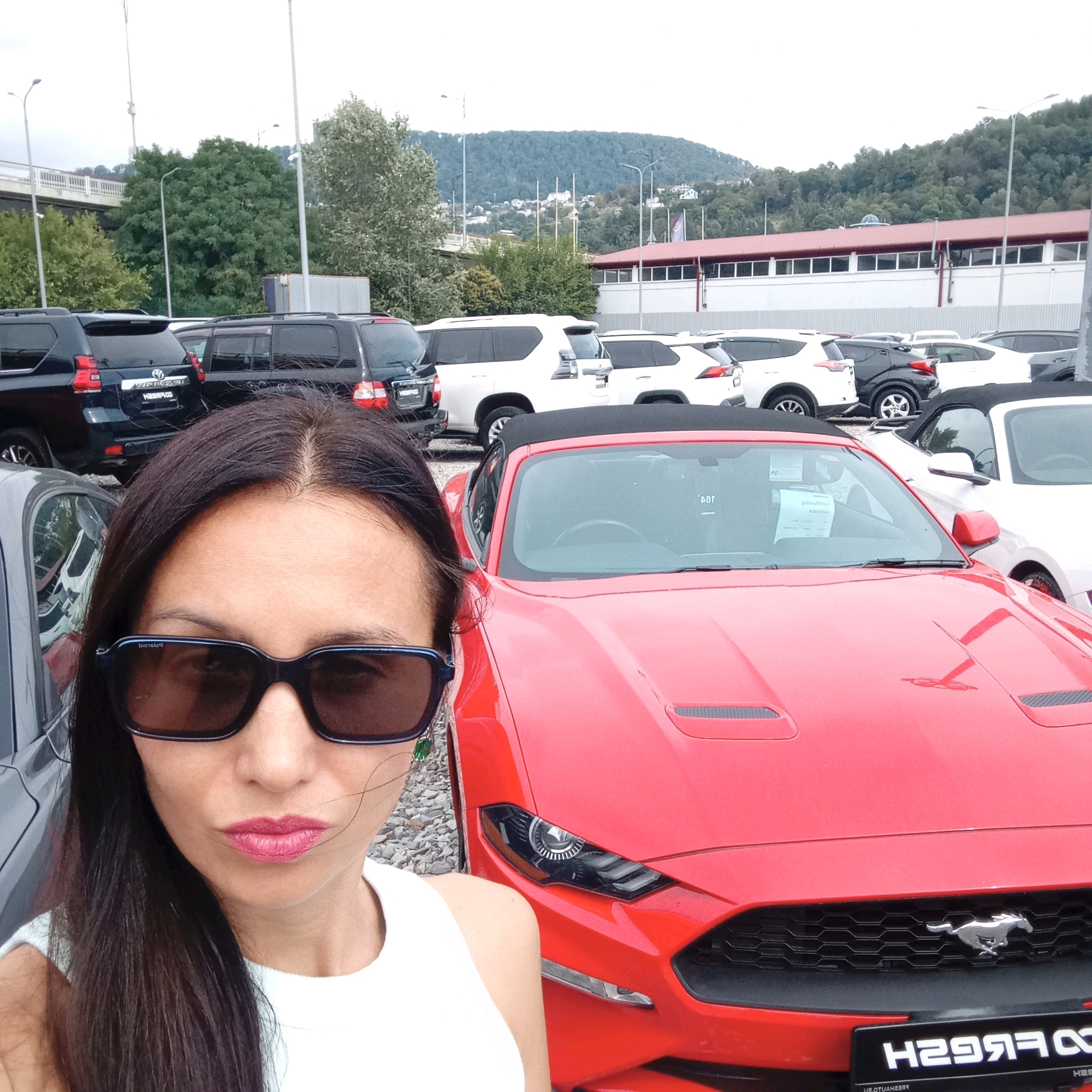 A selfie of a woman with dark hair and sunglasses is taken in front of a red Ford Mustang convertible. The woman is wearing a white tank top. In the background, there is a parking lot filled with various vehicles, including black SUVs and white cars. Beyond the cars, there is a building with a red roof and a white facade, and in the distance, a tree-covered hillside with some buildings is visible. The scene is outdoors on a cloudy day. The overall environment suggests a car dealership or a similar outdoor commercial space.