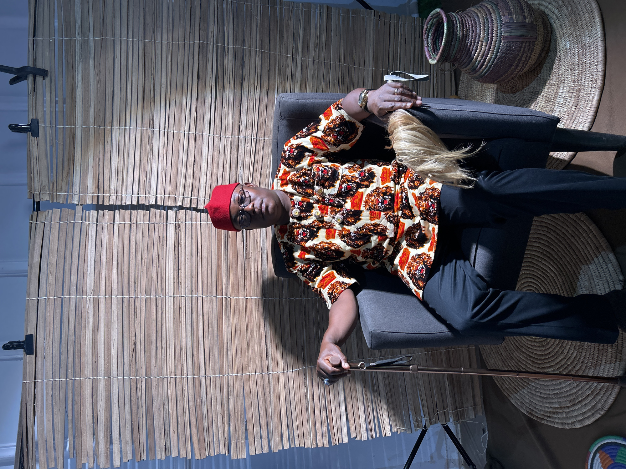 A man sits in a grey armchair in front of a woven bamboo backdrop and beside a large, round woven mat with a woven basket on top. The man wears a red cap, glasses, a patterned, button-down shirt with images of lion faces, and black pants. He is holding a silver object in one hand and a cane in the other. The scene appears to be staged in a studio or indoor setting. This image does not provide sufficient information to verify the location is in Jos, Nigeria.