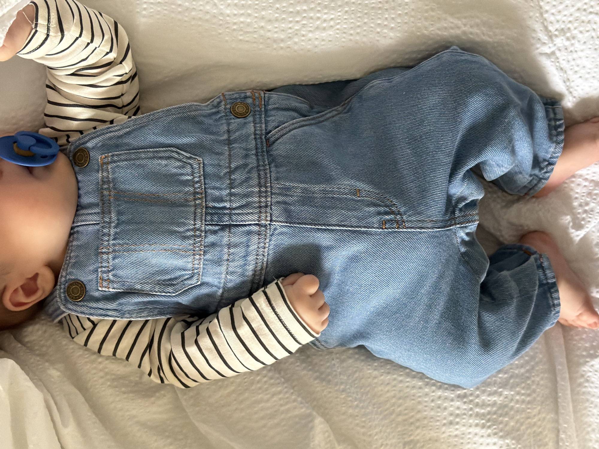 An overhead, slightly angled shot captures a baby lying on its back on a white, textured surface, likely a blanket or mattress cover. The baby's head is positioned to the left, with its bare feet visible to the right. A blue pacifier with a gold-colored accent is in the baby's mouth, suggesting a moment of rest or sleep.
The baby is dressed in a long-sleeved, cream or white shirt with thin black horizontal stripes, worn under light blue denim overalls. The overalls feature classic denim details, including brass-colored buttons, visible orange-brown stitching, and front pockets. Upon close inspection, some of the buttons bear faint text, with "MADE IN THE UK" and "ORIGINAL" being discernible.
Only parts of the baby's body are visible: its left ear, a cheek partially obscured by the pacifier, and its arms. One arm is bent with a small fist visible. The overall scene conveys a sense of calm and infancy, set indoors within a home environment, likely in Khimki, Russia. No specific time of day or external weather conditions can be determined from the image.