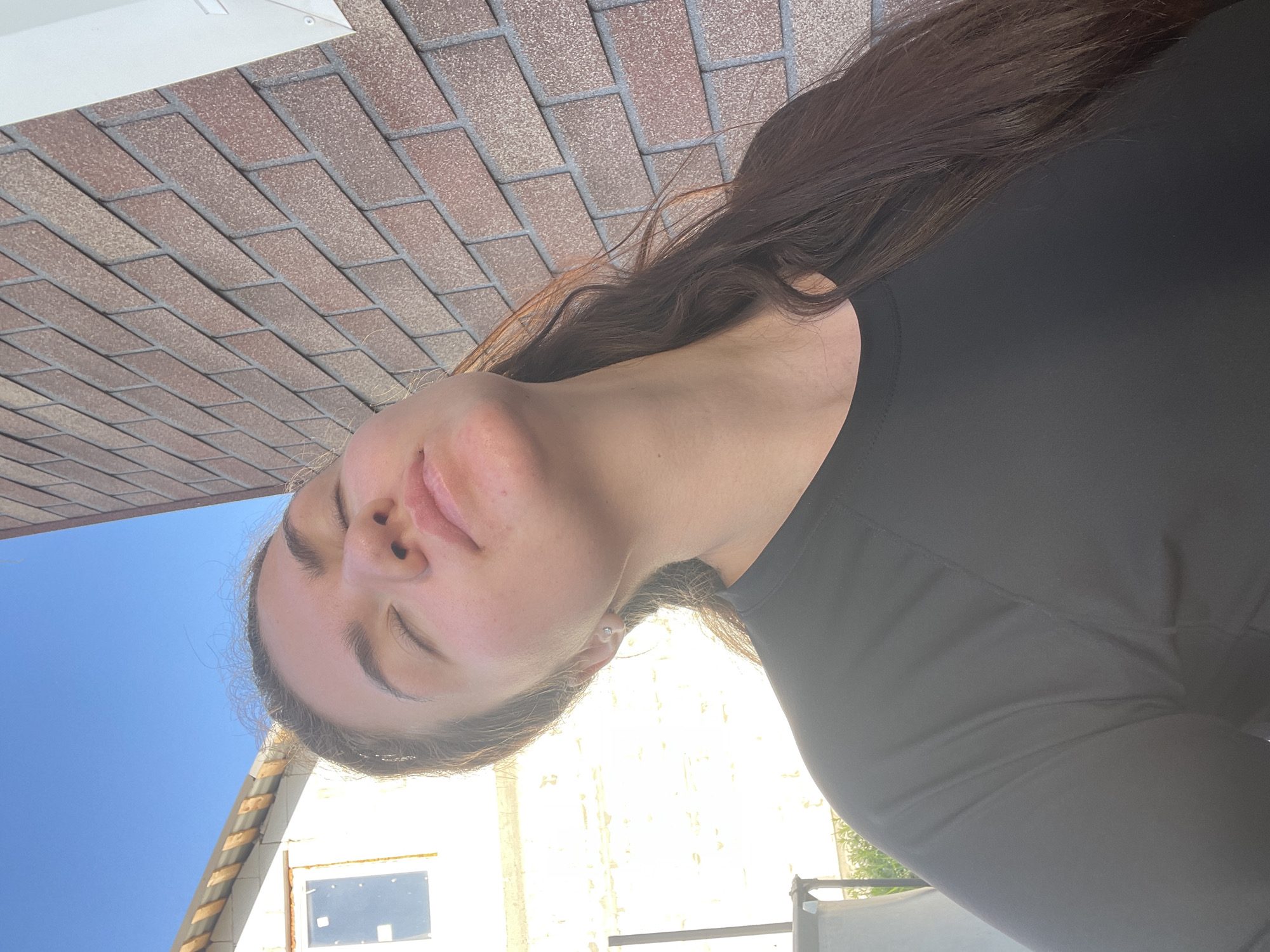A woman with dark hair is outdoors, viewed from a low angle. She wears a black long-sleeved shirt. Her eyes are closed. Behind her, in the background, are elements of the environment including a blue sky, the side of a building with a window, and brickwork, possibly a wall or another building element. The photograph appears to be taken from a street in Мосметаллоконструкция Онт, Russia, with the subject outside a building.