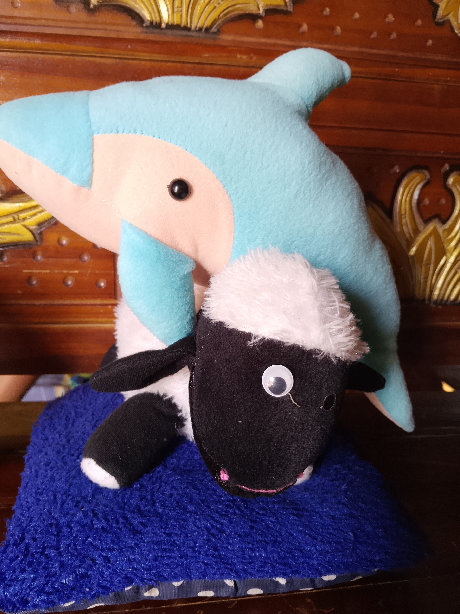 The image shows two stuffed toys on a wooden surface. The larger toy is a light blue shark with a peach-colored underbelly and a black eye. The smaller toy is a black sheep with white fluffy wool on its head and a single googly eye. The sheep is positioned in front of the shark, leaning slightly forward. Both toys are resting on a textured blue fabric, which is on top of a dark wooden surface. The wooden surface appears to be part of a piece of furniture, possibly a bench or table, with decorative golden trim. The background consists of more of the dark wooden furniture. The lighting suggests an indoor setting. There are no people visible in the image. The scene appears to be domestic. The location is specified as Kasiman, Indonesia.