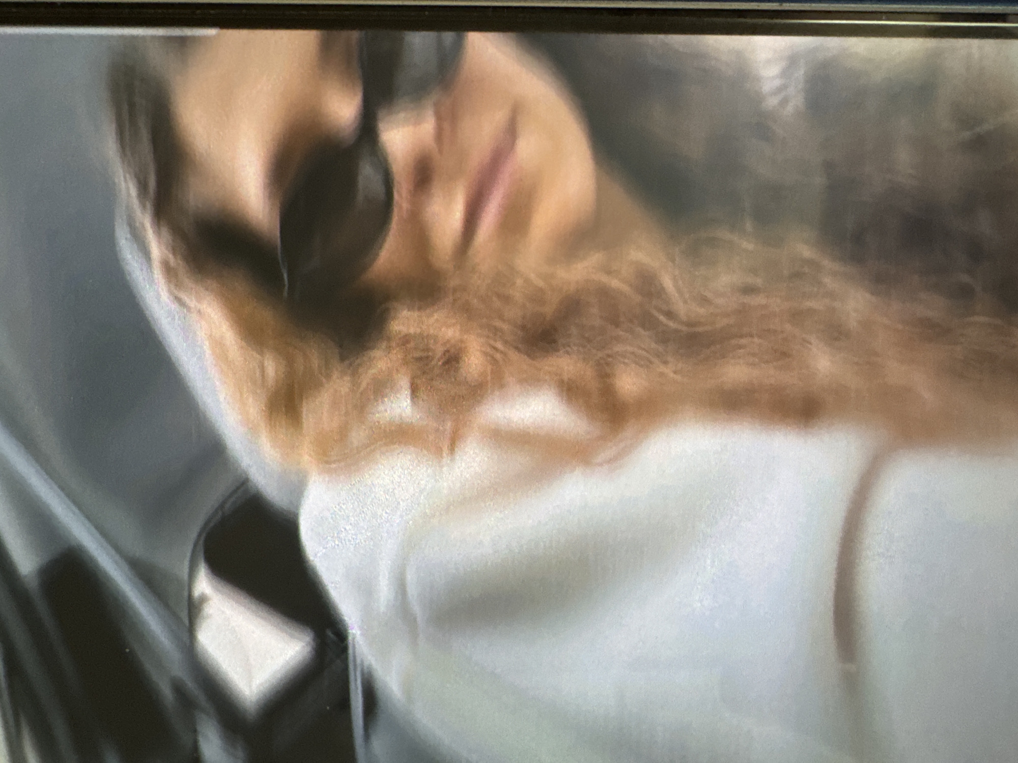 The image displays a blurry, vertically oriented, low-resolution photograph, potentially a screen capture or a photo of a screen, exhibiting significant motion blur and digital noise. A dark vertical bar is present on the far right edge of the frame.
The primary subject is an individual, likely female, positioned in the foreground, facing towards the right side of the frame with the head slightly tilted downwards. The person has long, wavy or curly hair, appearing in shades of auburn or light brown, visible cascading down the left side of the body and face. Dark, possibly black, sunglasses obscure the eyes and upper facial features. The lower facial area, including the nose, mouth, and chin, is visible but highly blurred, with the lips appearing slightly parted.
The individual is wearing a light-colored, possibly white or off-white, hooded garment, the fabric of which appears soft or fleecy due to the image artifacts. No specific details or logos are discernible on the clothing.
The background is dark and indistinct, suggesting an interior environment. Minimal details are identifiable, but vague reflections or highlights are present in the upper left area, possibly indicating a window or reflective surface. The image contains no discernible elements linking it to the city of Vratsa, Bulgaria.