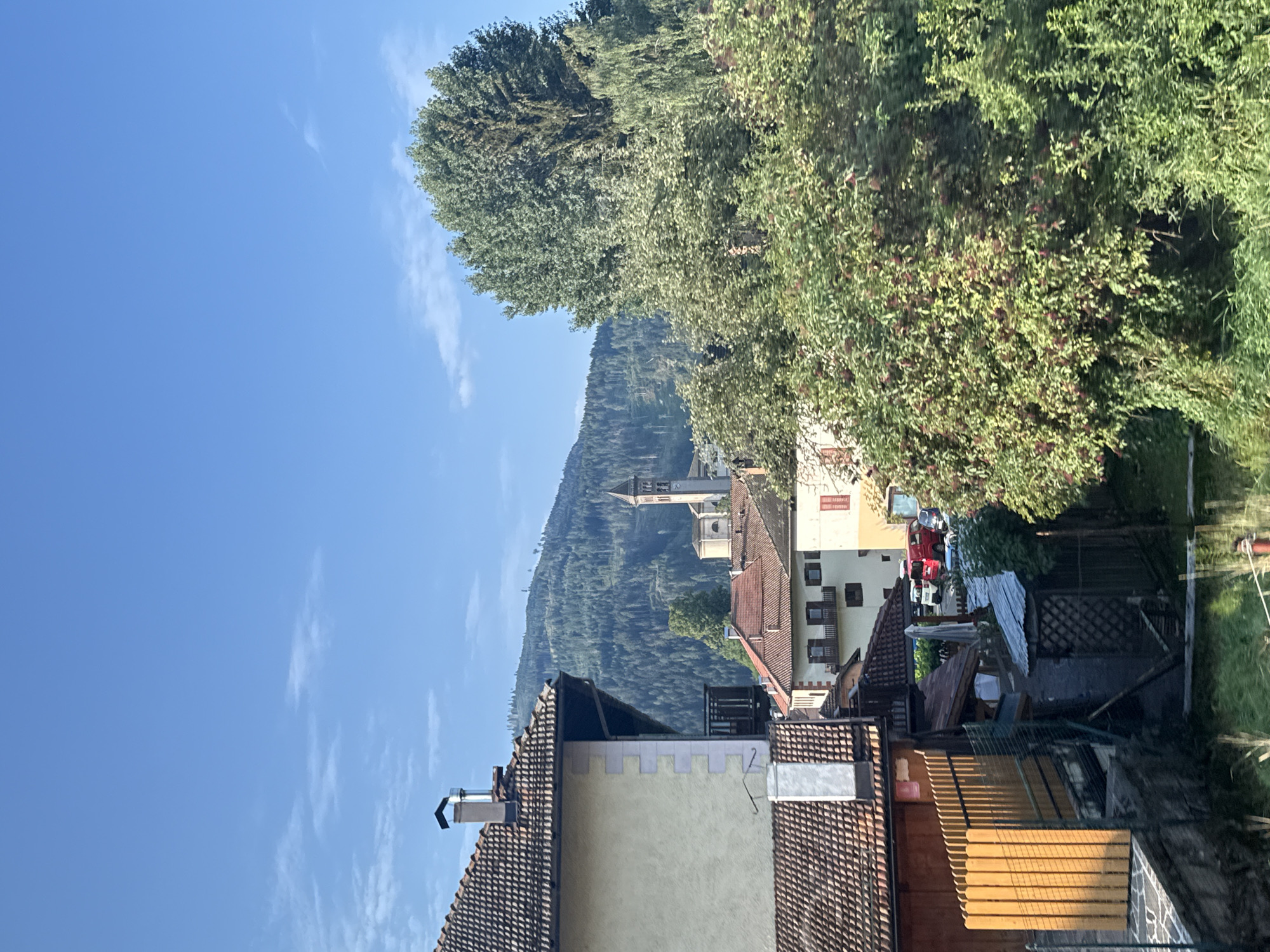 The image depicts a sunlit scene in Castello di Fiemme, Italy. In the foreground on the left, a building with a tiled roof and light-colored stucco walls is partially visible, with a wooden fence in front of it. Adjacent to this building, a smaller structure with a corrugated metal roof is seen. In the middle ground, several other buildings with red tiled roofs are clustered together, and a church steeple with a clock tower rises in the background. The background is dominated by a densely forested mountainside under a clear blue sky with scattered white clouds. The overall impression is of a peaceful, rural Italian town nestled amongst the mountains.
