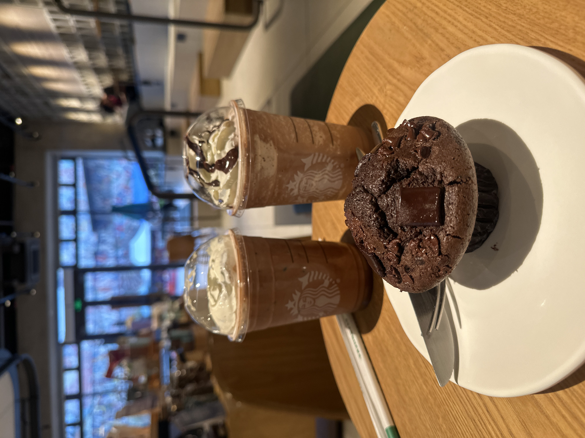 The image depicts a table in a Starbucks in Chongqing High-tech Zone, China. On the table are two Starbucks Frappuccinos, one with chocolate drizzle, and a chocolate muffin on a white plate. There's also a metal fork partially visible beneath the muffin and a Starbucks straw is also on the table. The background shows the interior of the Starbucks and a blurred view of the street outside.