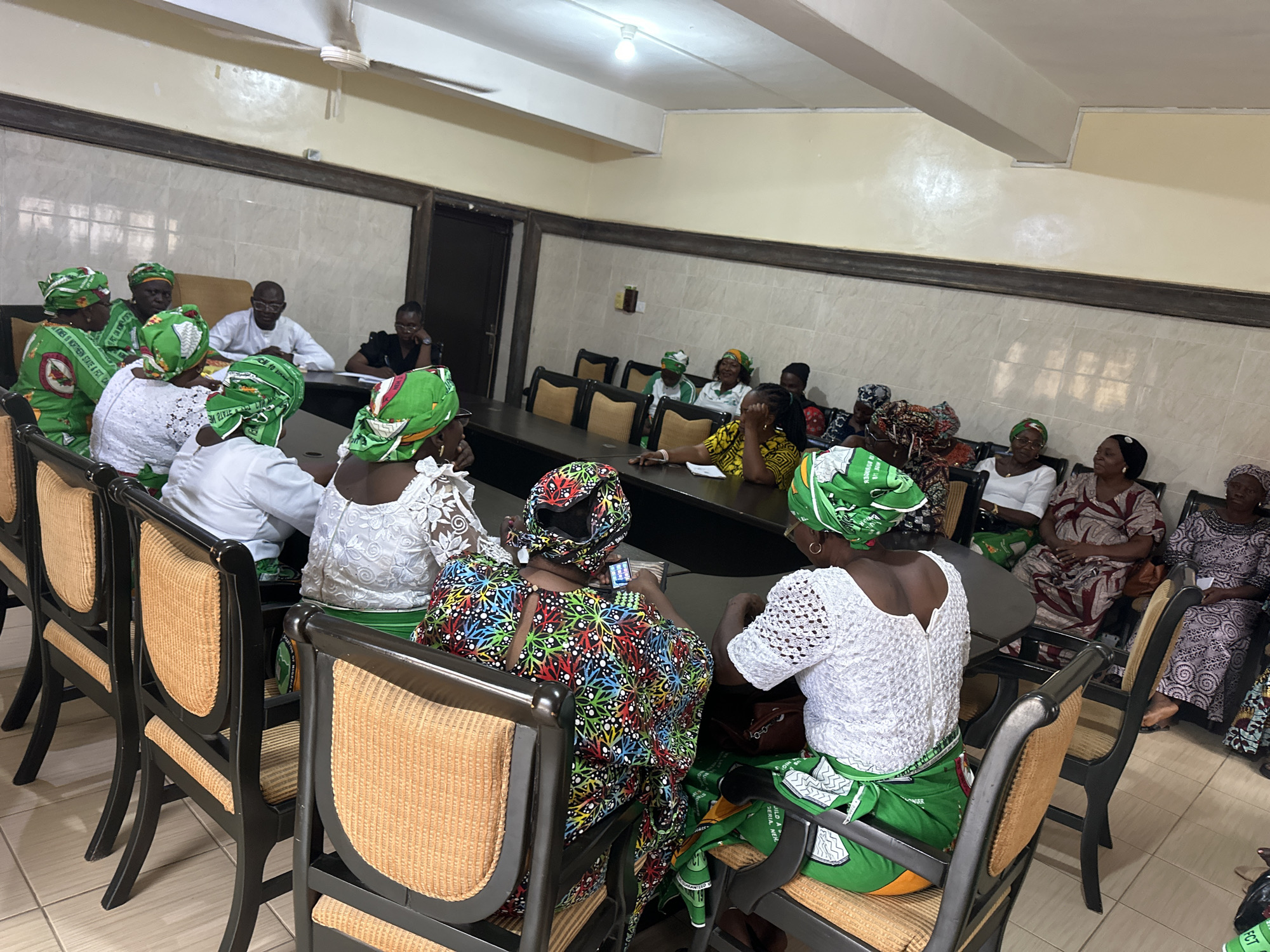 This media file depicts a group of people, predominantly women, gathered in a room that appears to be a meeting or conference space. The room has tiled walls and a beige ceiling with a light fixture and a ceiling fan. The subjects are seated around a large table, facing each other.
The women are dressed in vibrant, patterned traditional attire, including headwraps and long dresses or skirts. Many of the outfits feature green and white, some with elaborate designs and embroidered details. Some individuals are wearing glasses. There are a few men present, one of whom is wearing a white shirt and glasses, and another in the background wearing a dark shirt.
The activity appears to be a meeting or discussion, as individuals are looking towards each other and at the table. One woman in the foreground is looking down at a small electronic device, possibly a phone. The atmosphere seems engaged and possibly formal, given the setting and attire.
The lighting suggests it is daytime, with artificial lights on in the room. The tiled floor and the style of the chairs, which are dark wood with upholstered seats and backs, indicate an indoor setting. The overall impression is a communal gathering for discussion or a formal event.
There is some visible text on the green fabric worn by some of the women, appearing to be logos or inscriptions, though it is not clearly legible. The location context provided is Kabala, Nigeria, which aligns with the traditional attire and the style of the room.