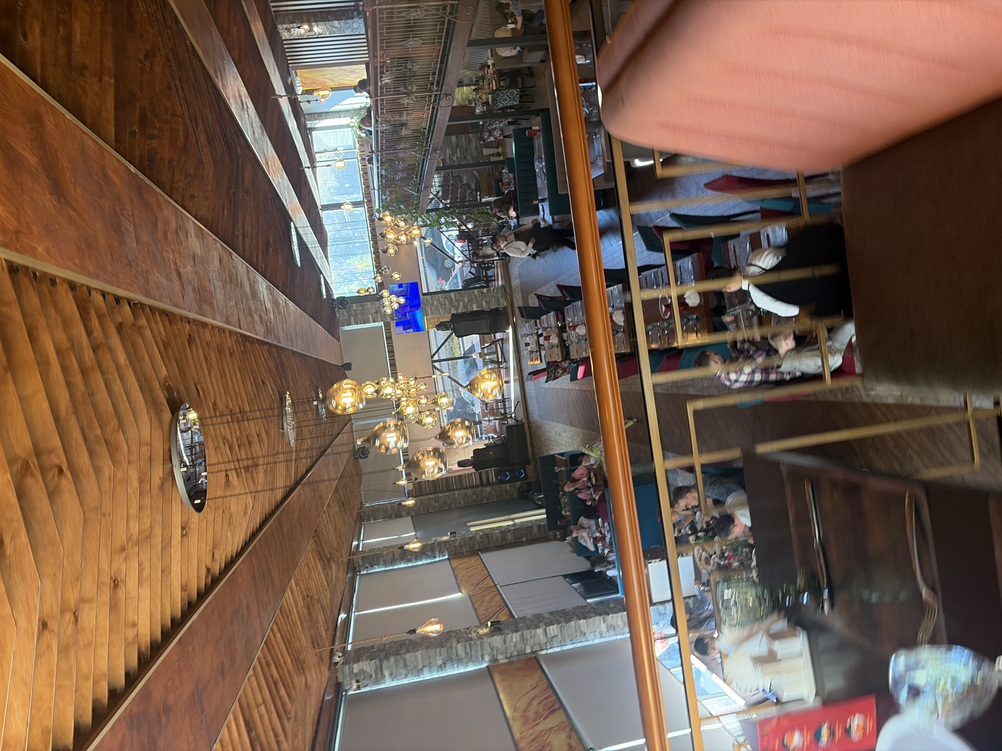 This image captures the interior of a multi-level restaurant or bar in Saint Petersburg, Russia, from a high vantage point, possibly from a table, with a person's hand visible in the upper right foreground.
The setting is an elegantly designed space featuring prominent wooden paneling on walls and ceilings, creating a warm, earthy ambiance. Large, modern chandeliers with numerous spherical light bulbs illuminate the different areas, complemented by natural light filtering in through expansive windows that reveal an outdoor view, possibly of trees or distant buildings. Sections of the walls are adorned with stone cladding, adding textural contrast.
The scene depicts a lively but not overcrowded atmosphere. Guests are seated at various tables across both the main floor and an elevated upper level, engaging in conversations and dining. At least one staff member, dressed in a black and white uniform, is visible on the lower level, attending to patrons. Reflections from a large mirror or polished surface on the right side of the frame offer a fragmented glimpse into other parts of the establishment, including more seating arrangements with colorful chairs and possibly additional patrons.
Notable details include circular decorative elements or small mirrors on the wooden wall panels, greenery placed along the railing of the upper level, and a large blue screen or television mounted on a pillar on the lower level. The presence of both natural and artificial light sources suggests it could be late afternoon or evening, typical for a dining venue. No discernible text is clearly visible within the frame.