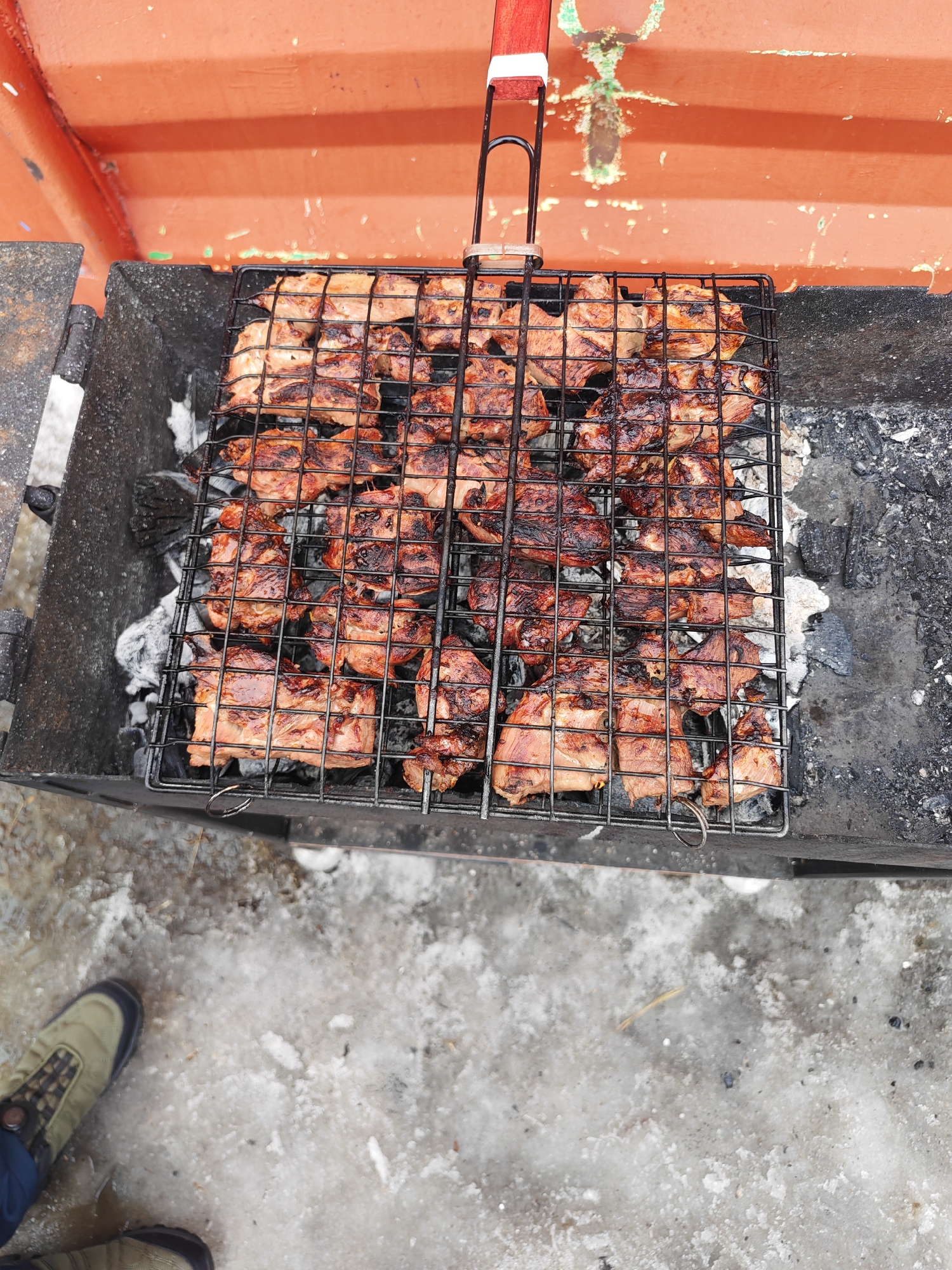 The image shows a charcoal grill filled with cuts of meat being cooked on a metal grate. The grill is situated outdoors, as evidenced by the presence of snow and ice on the ground. A person's shoe is visible in the lower-left corner of the frame. The grill is partially located in front of an orange structure. The setting appears to be in Ugolnye Kopi, Russia.