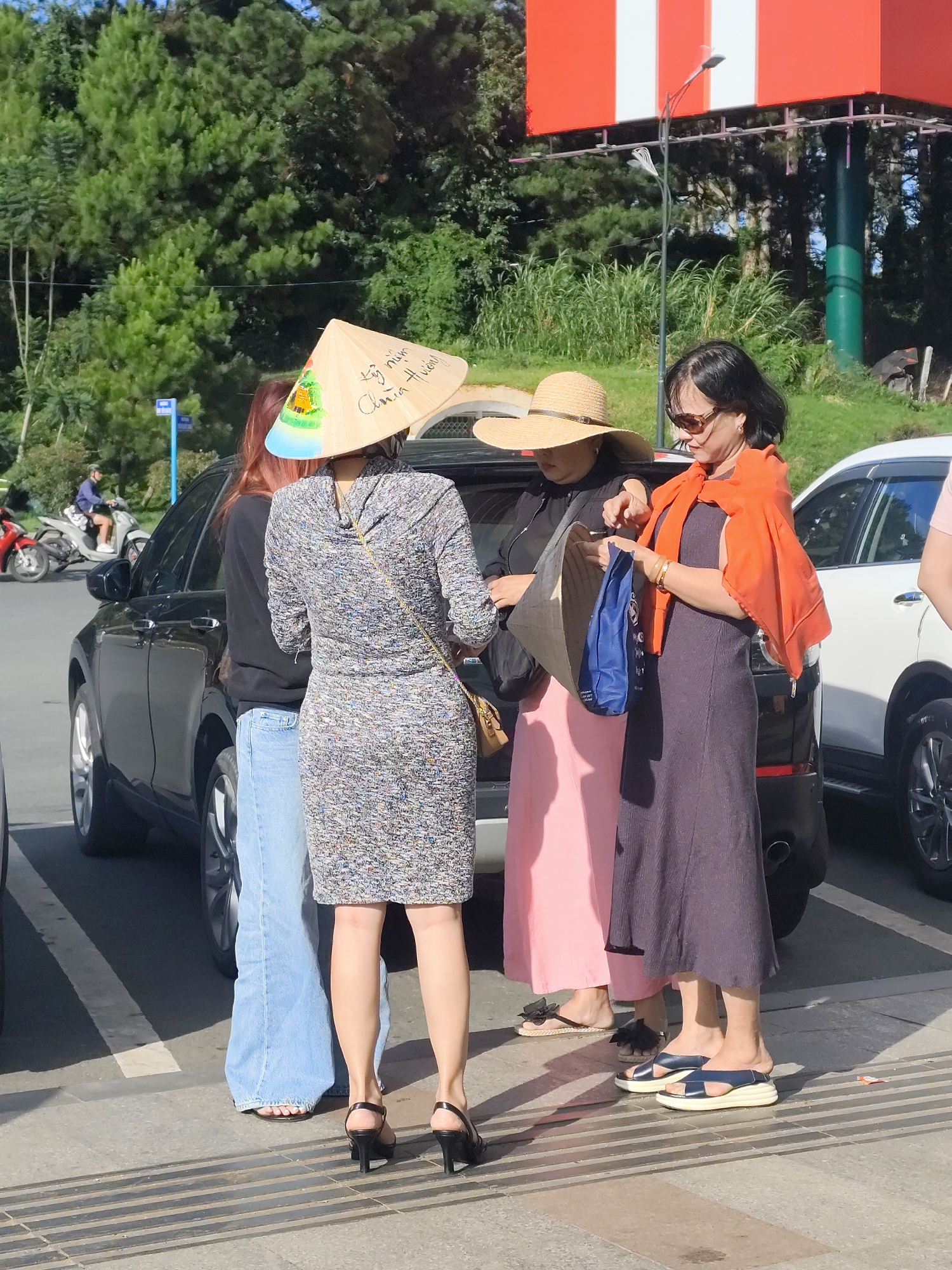 In Da Lat, Vietnam, four women are gathered near parked vehicles. One woman, on the left, wears a patterned dress and a conical hat. To her right, another woman is dressed in black with a wide-brimmed straw hat. She holds a conical hat and a blue bag. The third woman, in the center, wears a long pink skirt. The fourth woman, on the right, sports a dark dress and an orange shawl draped over her shoulders. She is holding a blue bag. The scene is set outdoors with trees and a billboard visible in the background. The ground is paved, and there are several cars present, including a black SUV and a white vehicle.