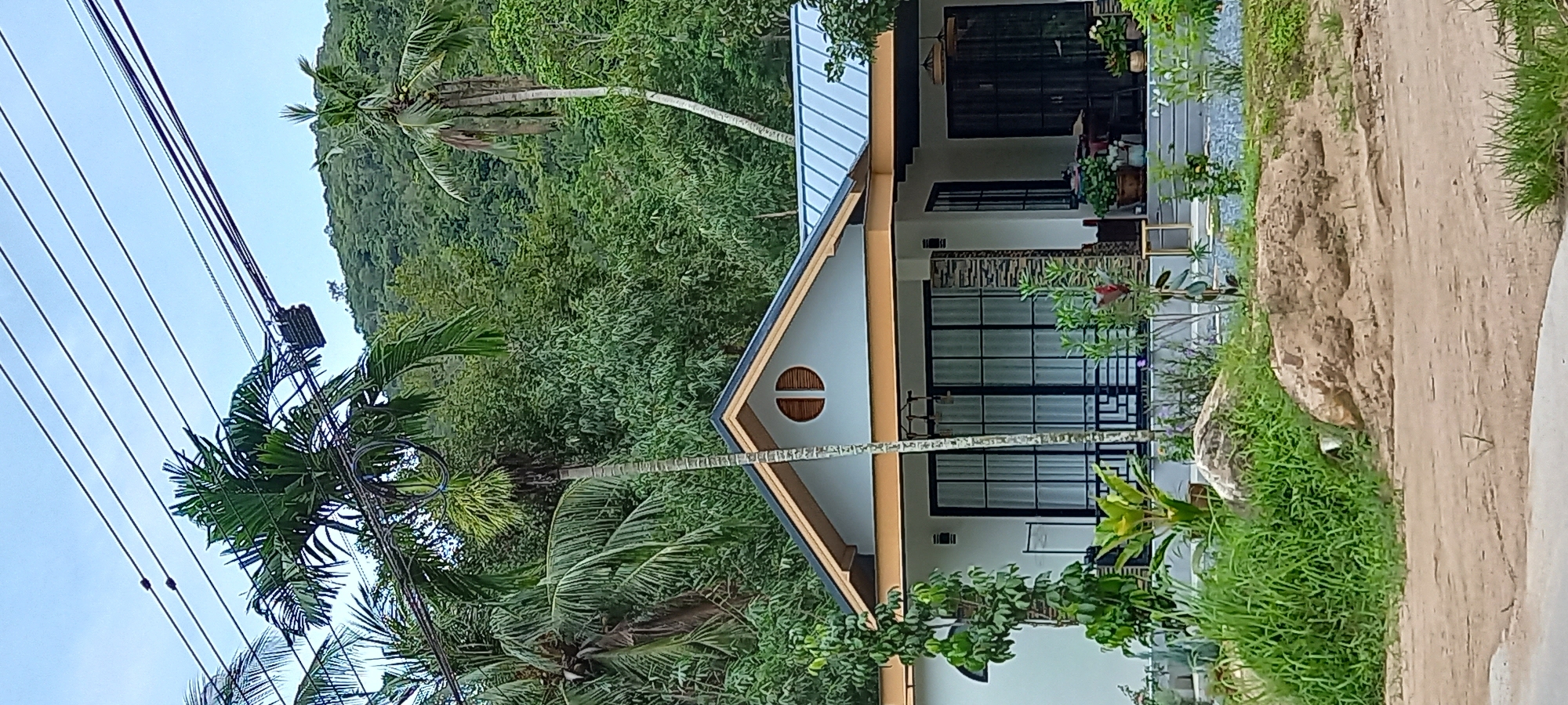 The image captures a view of a house nestled in a lush, green environment, likely located in the Tambon Koh Phangan area of Thailand. The house is a modern structure with a white exterior, black-framed windows, and a light blue metal roof. It features a distinctive circular window near the roof apex. Surrounding the house is dense foliage, with various trees and plants. A palm tree, visible on the left, is partially obscured by utility lines. To the right, a dirt path and more greenery suggest a natural setting. The overall scene conveys a serene and tranquil atmosphere, characteristic of a tropical island landscape.