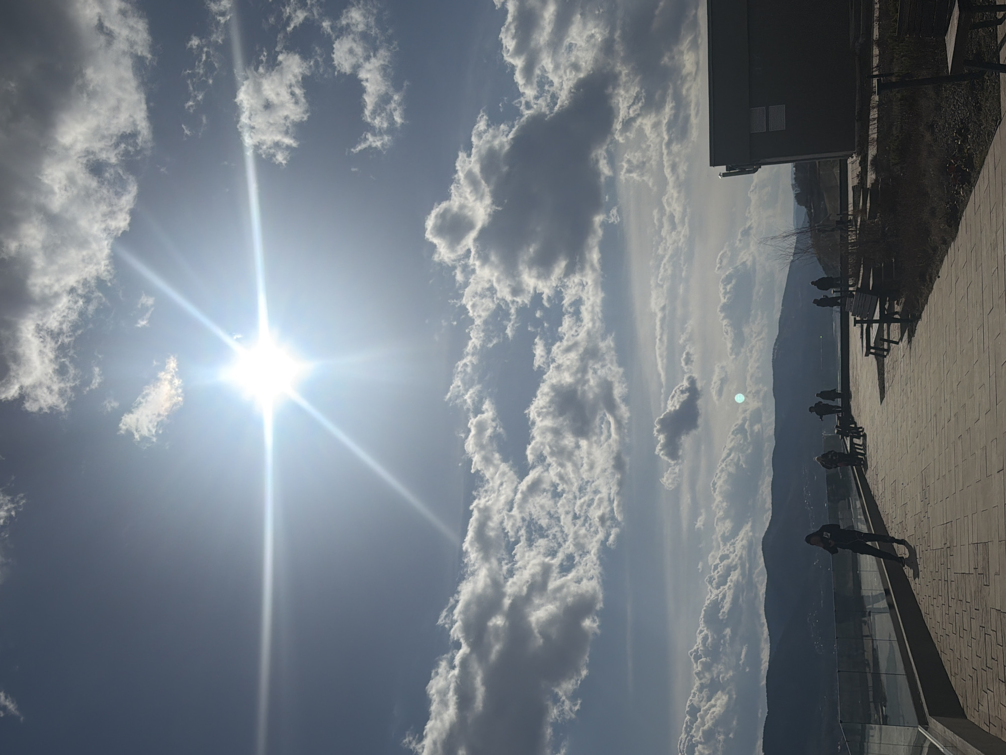 The image captures a sunny day in Krasnoyarsk, Russia, showcasing a bright sun piercing through a sky filled with scattered cumulus clouds. The camera appears to be positioned on an elevated walkway or terrace. Several people are visible along the terrace, some standing near the edge, looking out at the landscape, while others are seated on benches. A low mountain range is visible in the distance. The buildings and trees are on the right.