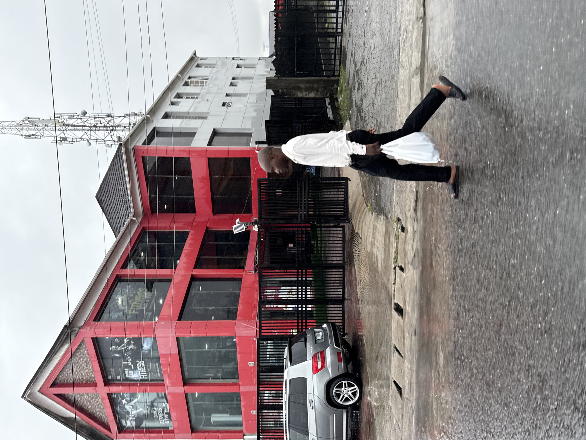 The image shows a street scene in Ado, Nigeria, on a wet day. A man dressed in a white shirt and black pants is walking on the sidewalk, carrying a white plastic bag. He is moving from right to left in the frame.
Behind him is a multi-story building with a red facade and large windows. The building appears to be a fitness center or gym, as indicated by the signage "DOL FITNESS GYM" visible on the ground floor. There is a silver SUV parked in front of the building.
To the right of the building is a section of black metal fence and some green vegetation. Further in the background, there is another light-colored building and a communication tower with antennas. The street is wet, suggesting recent rain. The sky is overcast.