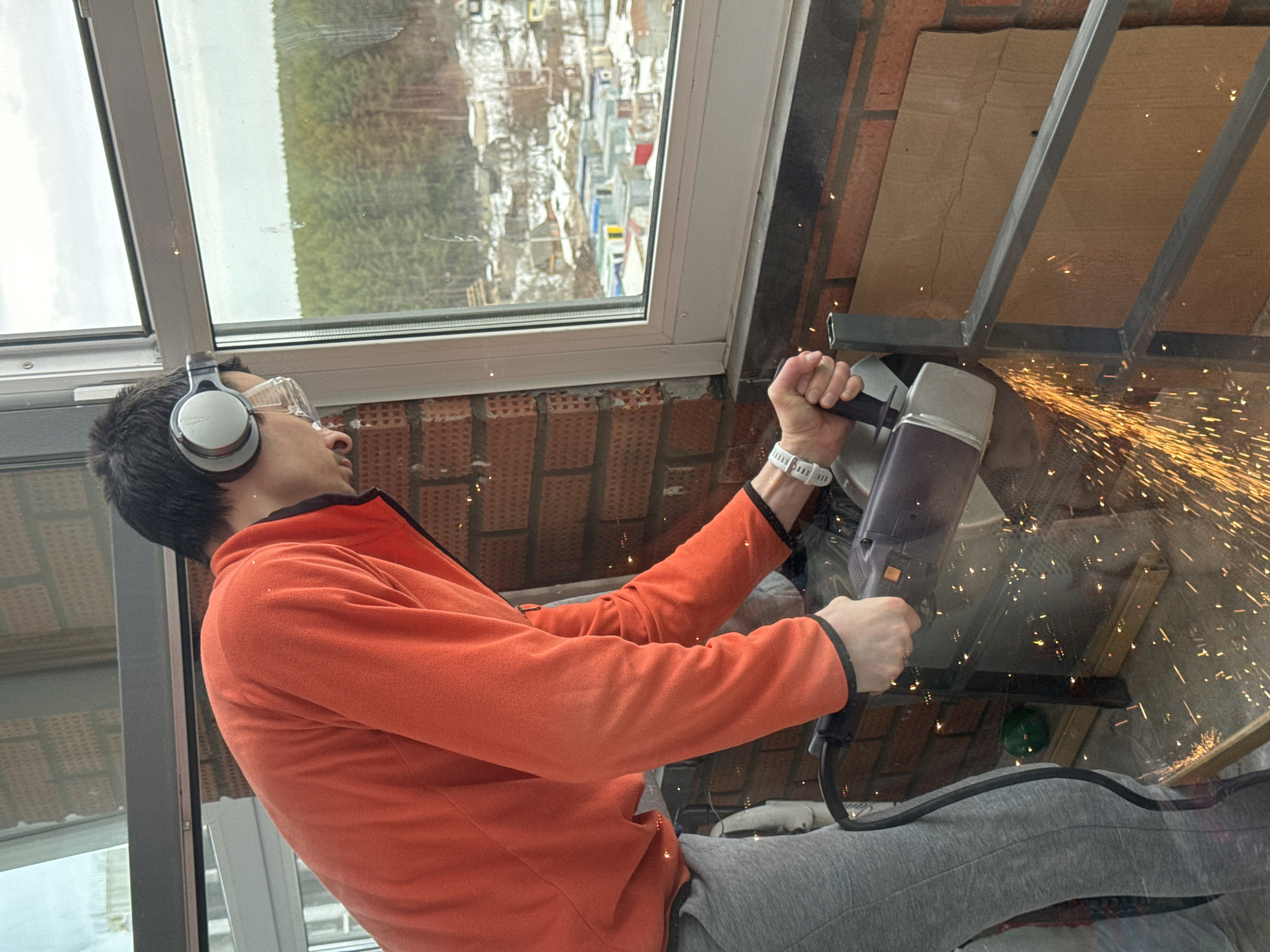 A man is cutting metal with a power tool indoors, producing sparks. He is wearing safety glasses and headphones. He has on an orange sweater and gray sweatpants. A window reveals a view of a city with trees in the distance. Based on the distinctive urban landscape, the city is likely Izhevsk, Russia. The man is cutting a metal frame on top of cardboard. The wall is brick.