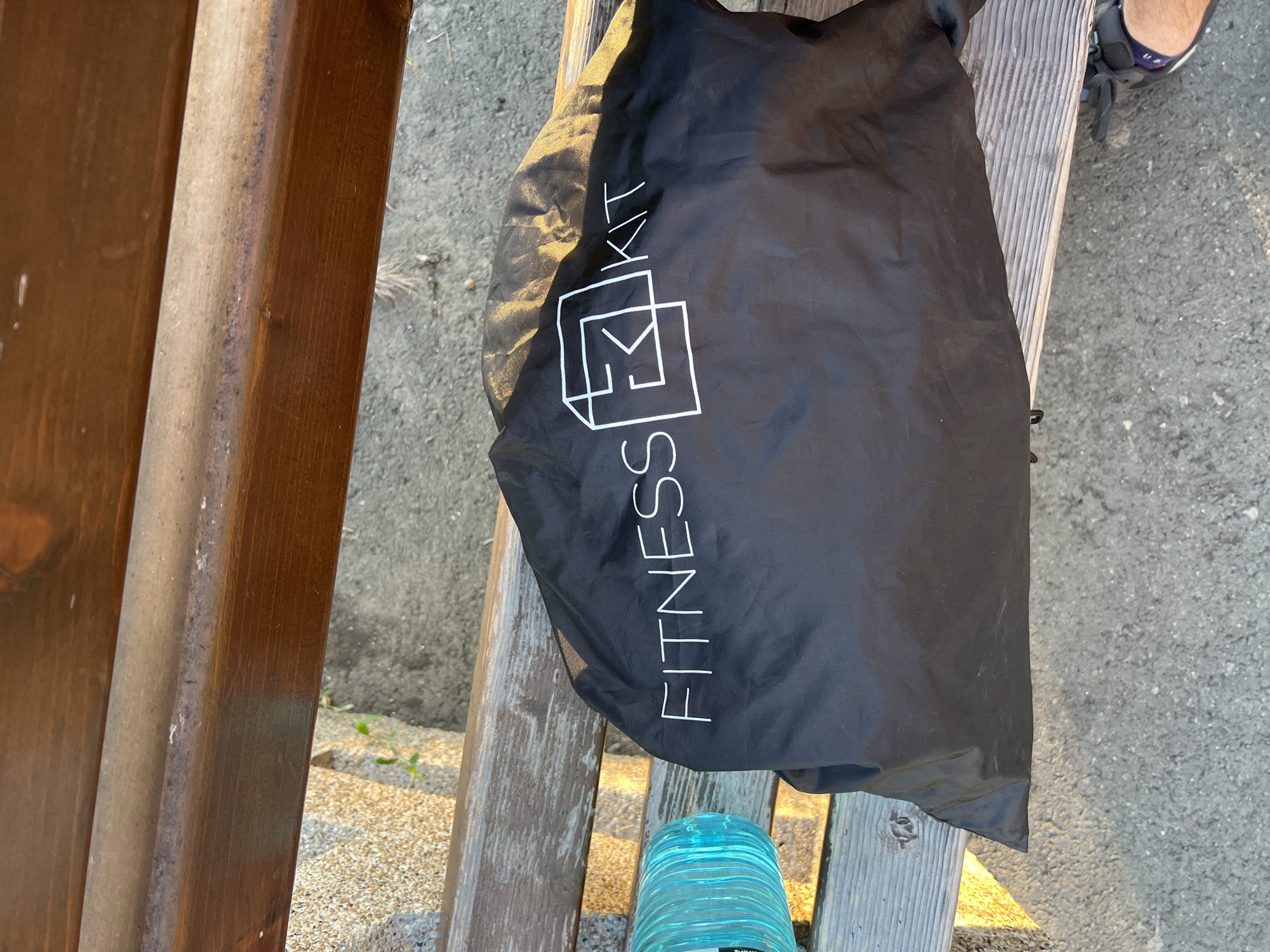 A black fabric bag with white text "FITNESS KIT" and a square logo is placed on a wooden bench. A turquoise plastic water bottle is to the left of the bag. The bench is positioned outdoors on a surface that appears to be asphalt or concrete. A person's foot wearing a sandal is visible at the bottom right of the image. The scene is illuminated by sunlight casting shadows. The background shows a sandy area with some dry vegetation. The location is an outdoor seating area in Burgas, Bulgaria.