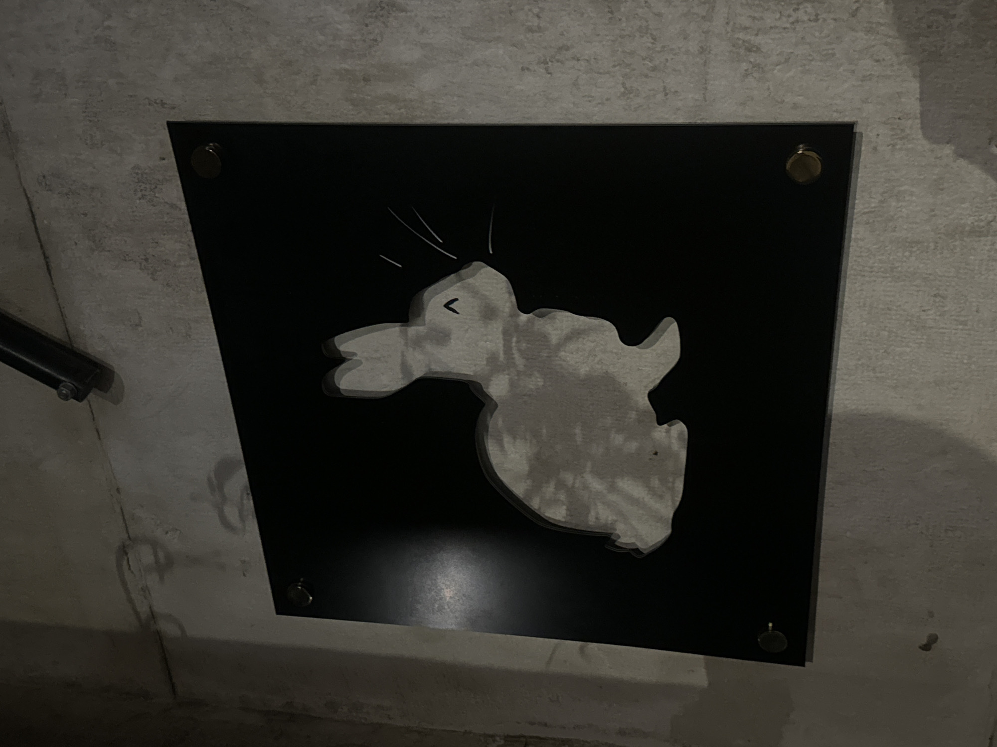 A black, square-shaped plaque featuring a stylized white silhouette of a rabbit is mounted on a light gray, textured concrete wall in Sofia, Bulgaria. The rabbit, depicted in profile facing right, has upright ears, a small "^" shape for its nose/mouth, and three white lines indicating whiskers. The white rabbit silhouette has a subtle shadow effect, creating a perception of depth against the plaque's matte black surface. The plaque is secured to the wall with four visible brass or gold-colored standoffs, one positioned in each corner. The concrete wall displays variations in its light gray tone and texture, with some faint, irregular darker markings visible, predominantly above the plaque. A dark, cylindrical object, possibly part of a fixture, is partially visible at the upper left edge of the frame. The lighting in the scene is dim, with a minor reflection present on the left side of the black plaque. No people or active interactions are visible in this interior setting.