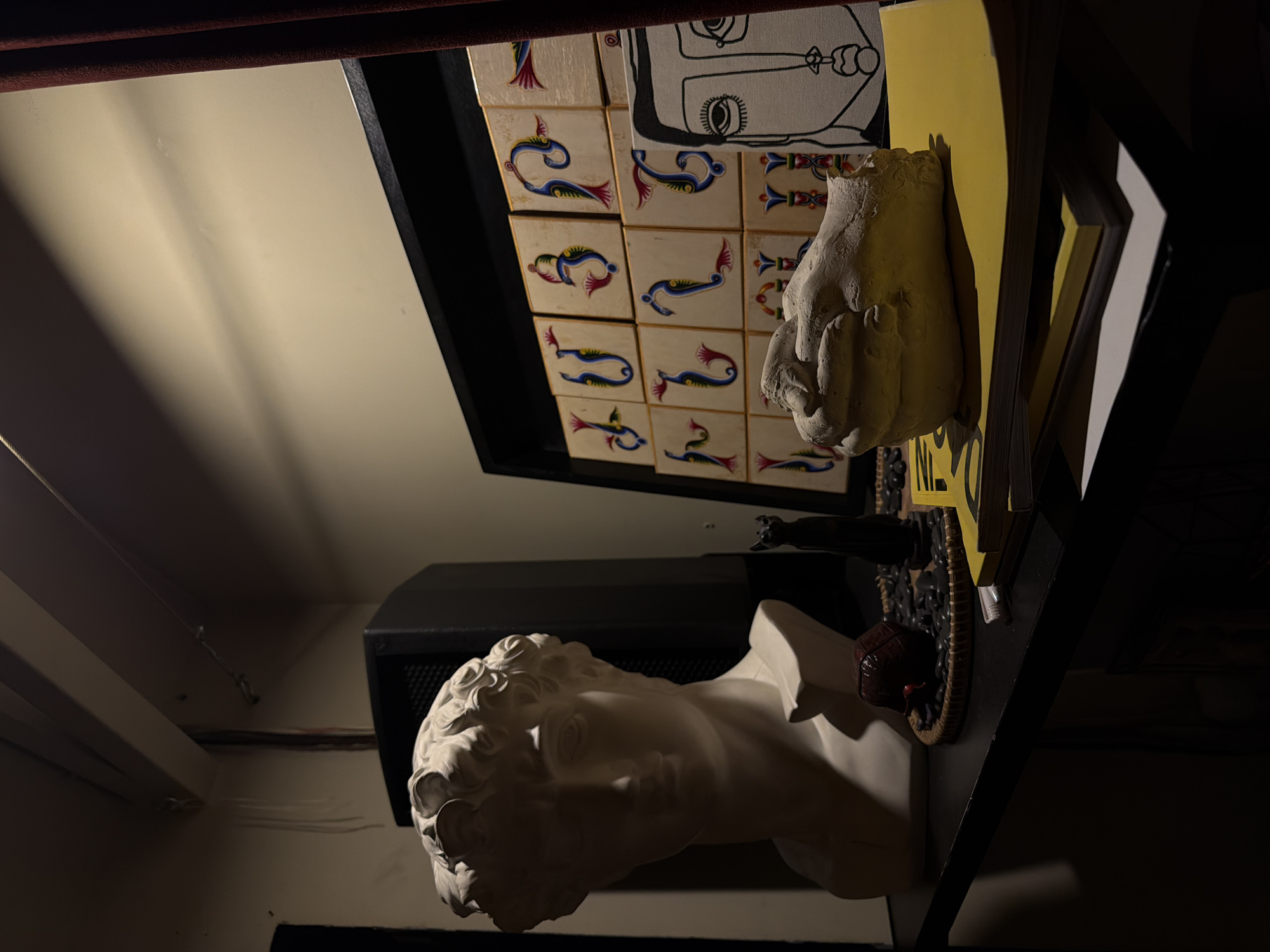 A low-angle indoor shot depicts a collection of objects on shelves. The objects include a bust of David's head, a bust of a hand, painted tiles, and yellow papers. The shot is taken in Yerevan, Armenia.