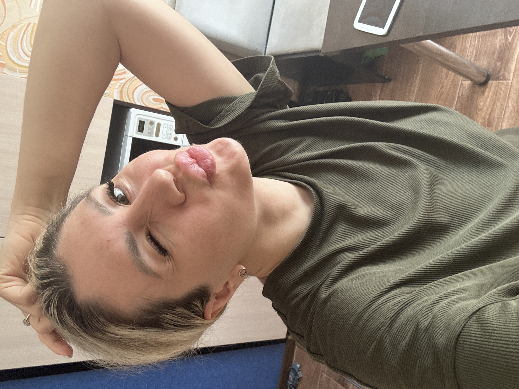 A woman with blonde hair pulled back and a ring on her right hand is in the foreground, winking and puckering her lips at the camera. She is wearing a green, ribbed t-shirt. Behind her, to the left, is a blue wall. To the right is a microwave oven and a cabinet. Further right, a dark wooden table with a tablet on it is visible, and part of a wooden chair leg. The flooring appears to be wood. The scene is likely indoors, in a residential setting, possibly a kitchen or dining area. There is no indication that this location is in Novoaltaysk, Russia.