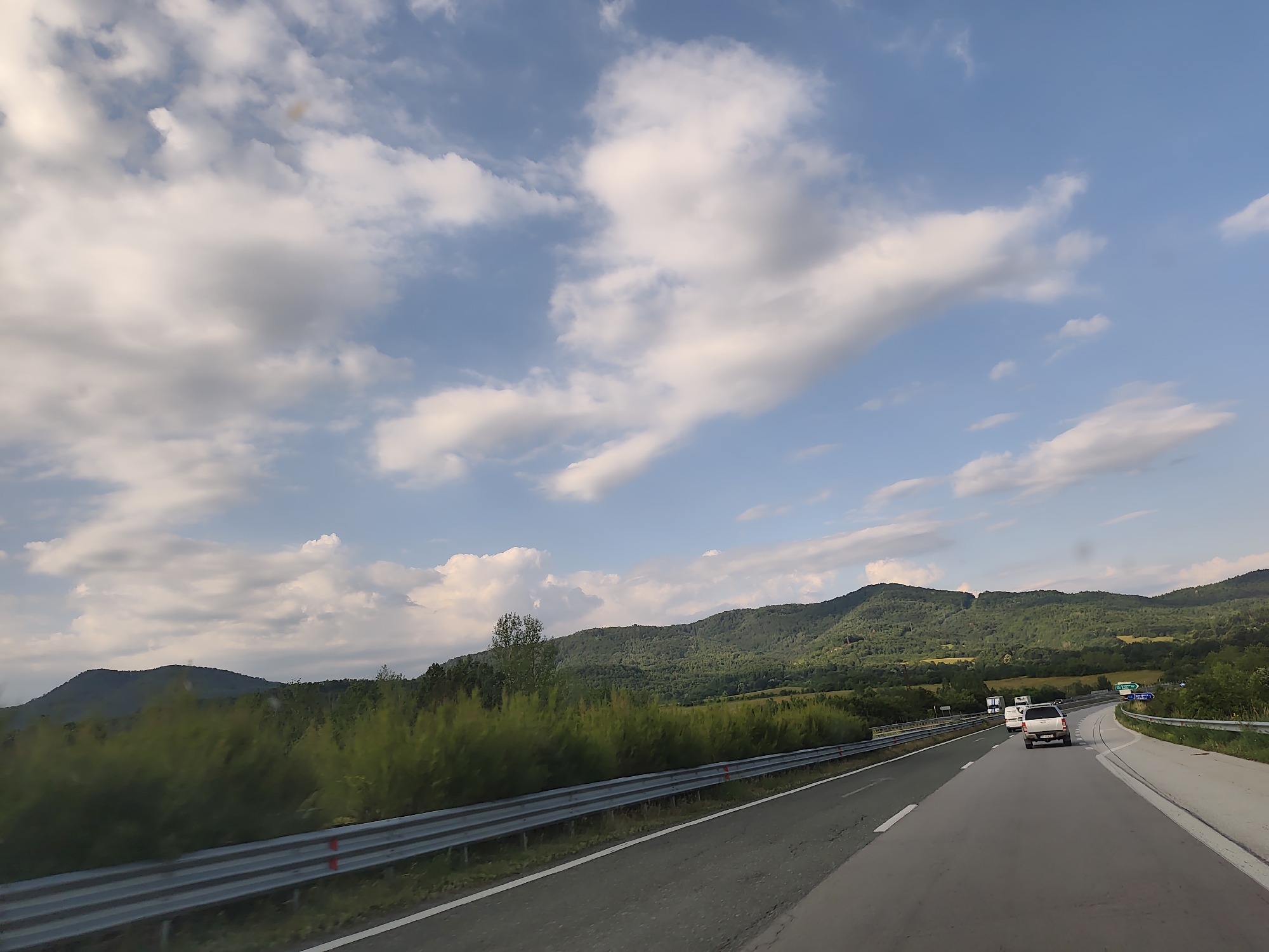 The image shows a highway with a metallic guardrail on the left. There are trees and rolling hills in the background. The sky is blue with scattered clouds. Two vehicles, a white van and a white SUV, are traveling on the highway. The scene appears to be in a rural or mountainous area.