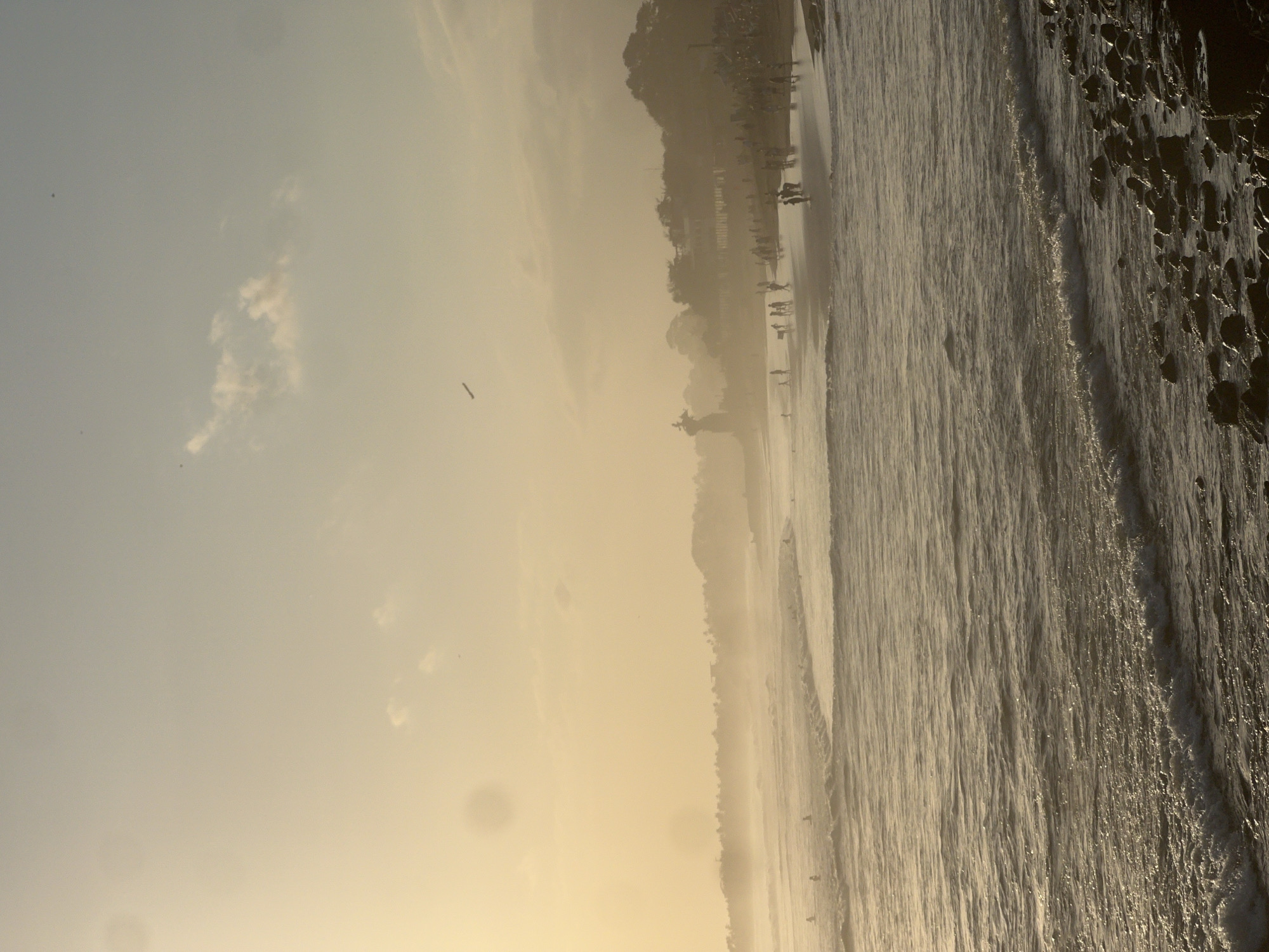 The image captures a coastal scene at sunset in North Kuta, Indonesia. The sky is a hazy yellow, with soft clouds and a distant airplane visible. The ocean has moderate waves, with some people visible in the water, possibly surfing or swimming. The shoreline is populated with numerous individuals walking along the beach or gathered in groups. A prominent statue or monument stands on the left side of the beach, silhouetted against the hazy background. Lush vegetation lines the coastline, with trees and buildings partially obscured by the atmospheric conditions. The foreground features a rocky shore with small waves lapping at the stones. The overall impression is one of a serene, yet active, beach at dusk.