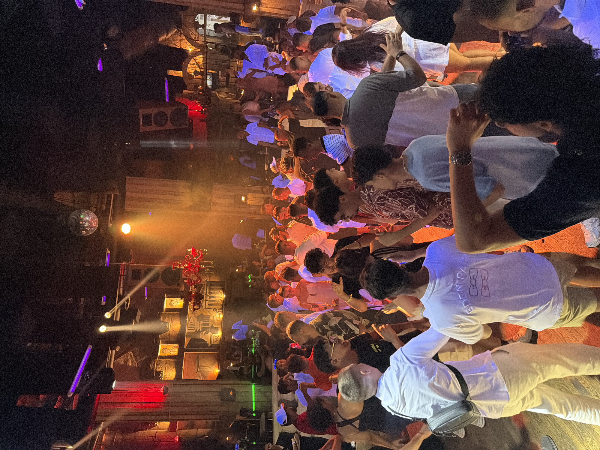 The image depicts a crowded indoor nightclub scene in Sharm El Sheikh, Egypt, characterized by dim, atmospheric lighting from stage lights and a disco ball overhead. A diverse group of approximately 50-70 individuals, predominantly young adults, are visible, engaged in dancing and socializing. The architecture features ornate detailing, including columns and wall reliefs reminiscent of ancient Egyptian or Greco-Roman styles, suggesting a themed or upscale venue. Red and orange lighting casts a warm glow across the scene, highlighting the vibrant energy of the partygoers. The attire of the attendees ranges from casual to semi-formal, with many wearing light-colored clothing suitable for warm weather. Several individuals are captured mid-movement, their gestures indicating active participation in dancing. The overall impression is one of a lively and energetic social gathering at a popular entertainment establishment.
