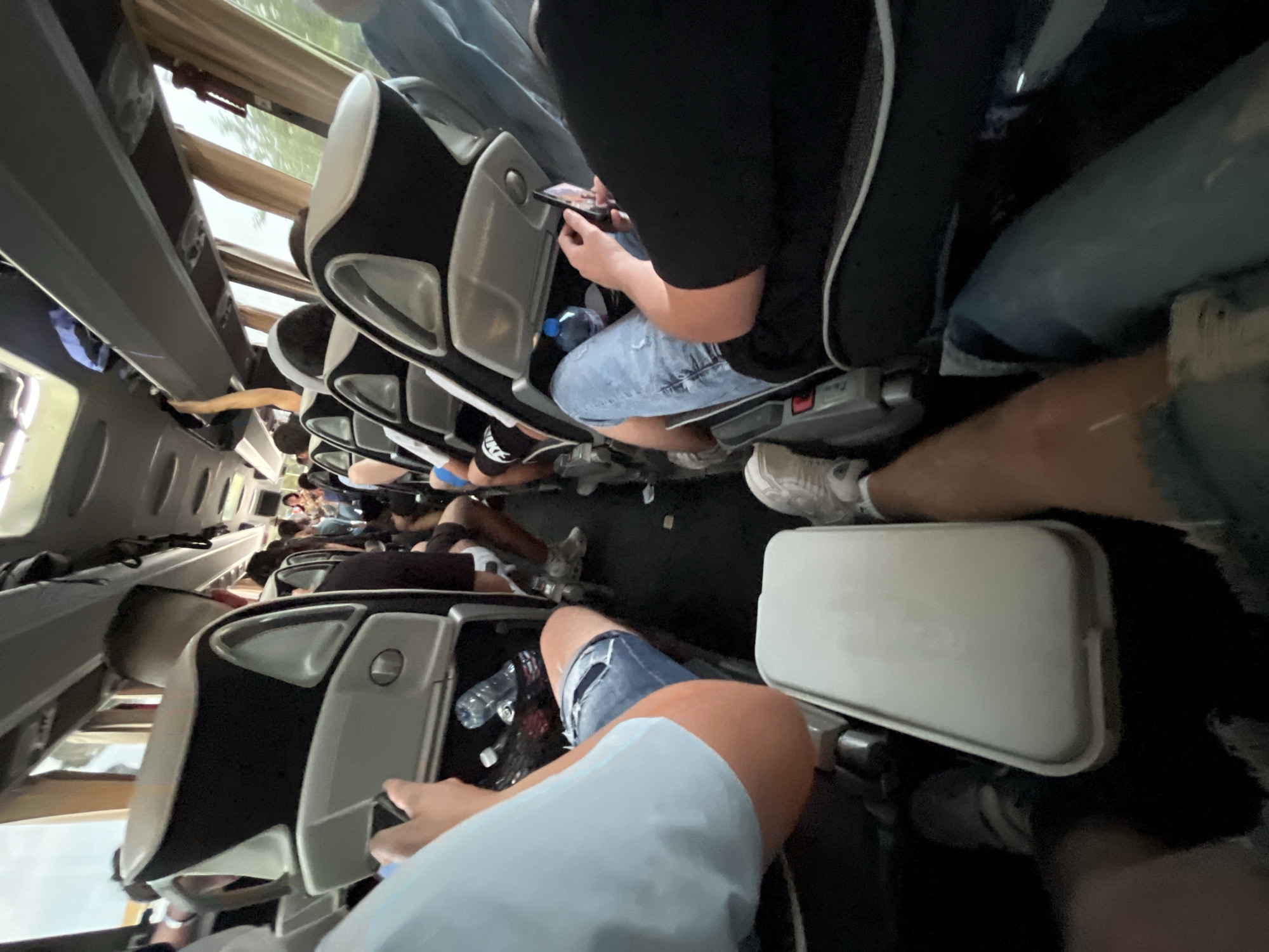 The image depicts the interior of a bus filled with passengers. The bus is likely in motion, as suggested by the blurred background seen through the windows. Passengers are seated in black and gray seats. People are of various ages, and wear clothes such as t-shirts and jeans. One passenger is using a smartphone. Water bottles can be seen in the seat pockets in front. Debris is present on the floor, suggesting the bus is likely being used for long journeys. The location of the bus in the city of Ca' Savio, Italy, is not discernible from the visual information.