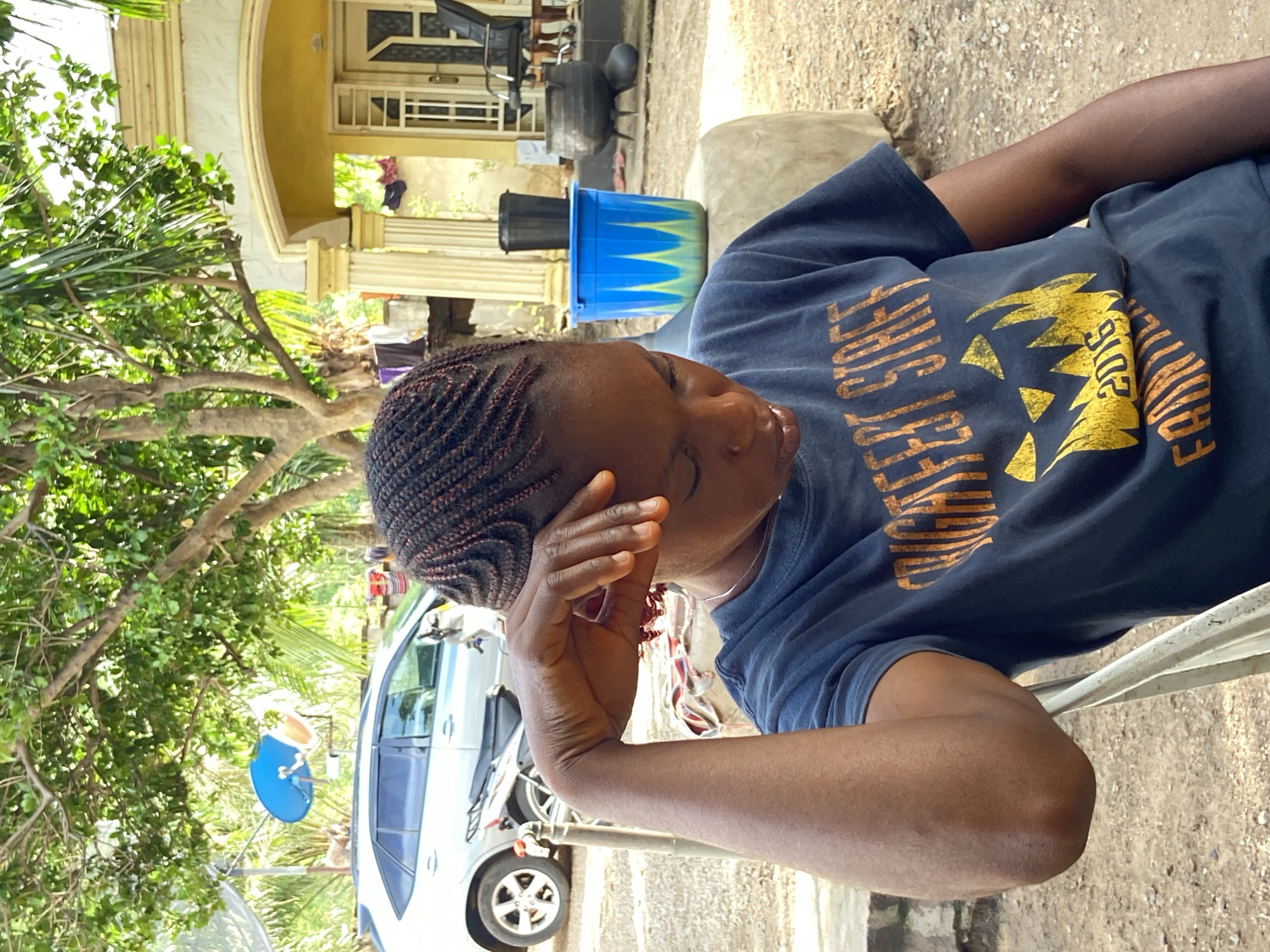 The scene is an outdoor setting in Mararaba, Nigeria. A dark-skinned woman with braided hair is sitting on a metal chair. She wears a navy-blue T-shirt with gold text and graphic. Her right hand is near her face. Behind her is a residential building with yellow accents. To the left is a car, partially obscured by a tree with lush green leaves. A blue satellite dish is also visible near the car. A blue and green bucket and a dark-colored trash can are behind the woman. The ground appears to be dirt and gravel.