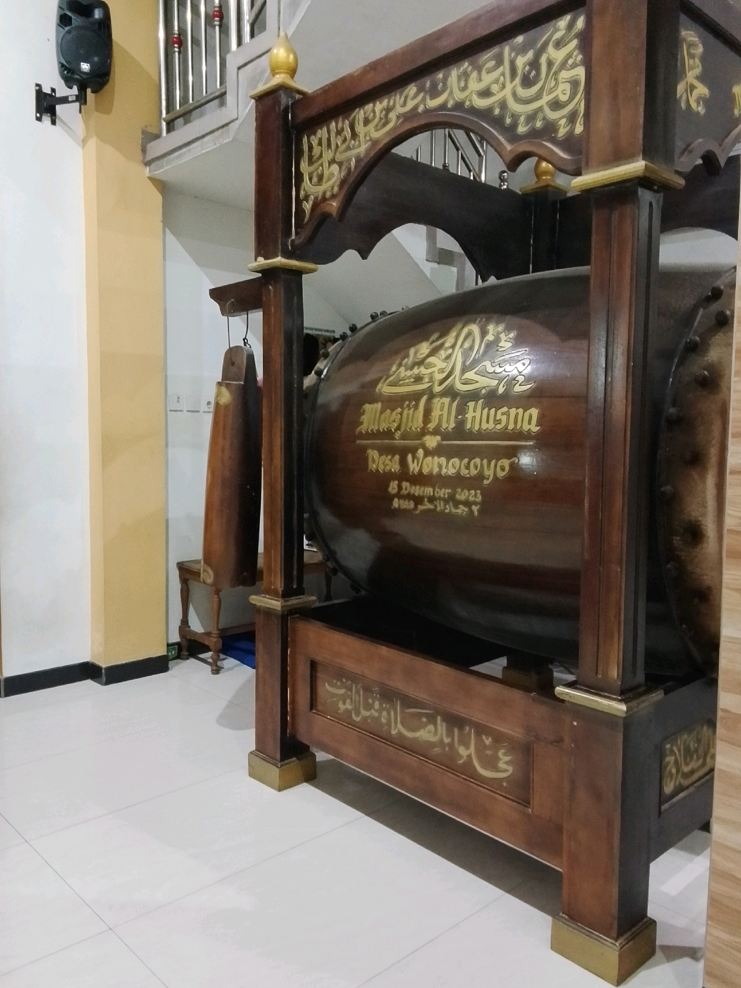 The image shows a large, wooden drum, likely a mosque drum (bedug), adorned with gold calligraphy. The drum is mounted on an ornate wooden frame with further gold inscriptions. Text on the drum reads "Masjid Al-Husna, Desa Wonocoyo, 15 Desember 2023". Hanging to the left of the drum is a wooden percussion instrument, possibly a kentongan. The drum and frame are set against a light-colored wall and tiled floor. In the background, a staircase with metal railings is visible. To the left, a black speaker is mounted on the wall. The scene appears to be indoors, within or near a building in the village of Wonocoyo, Indonesia.
