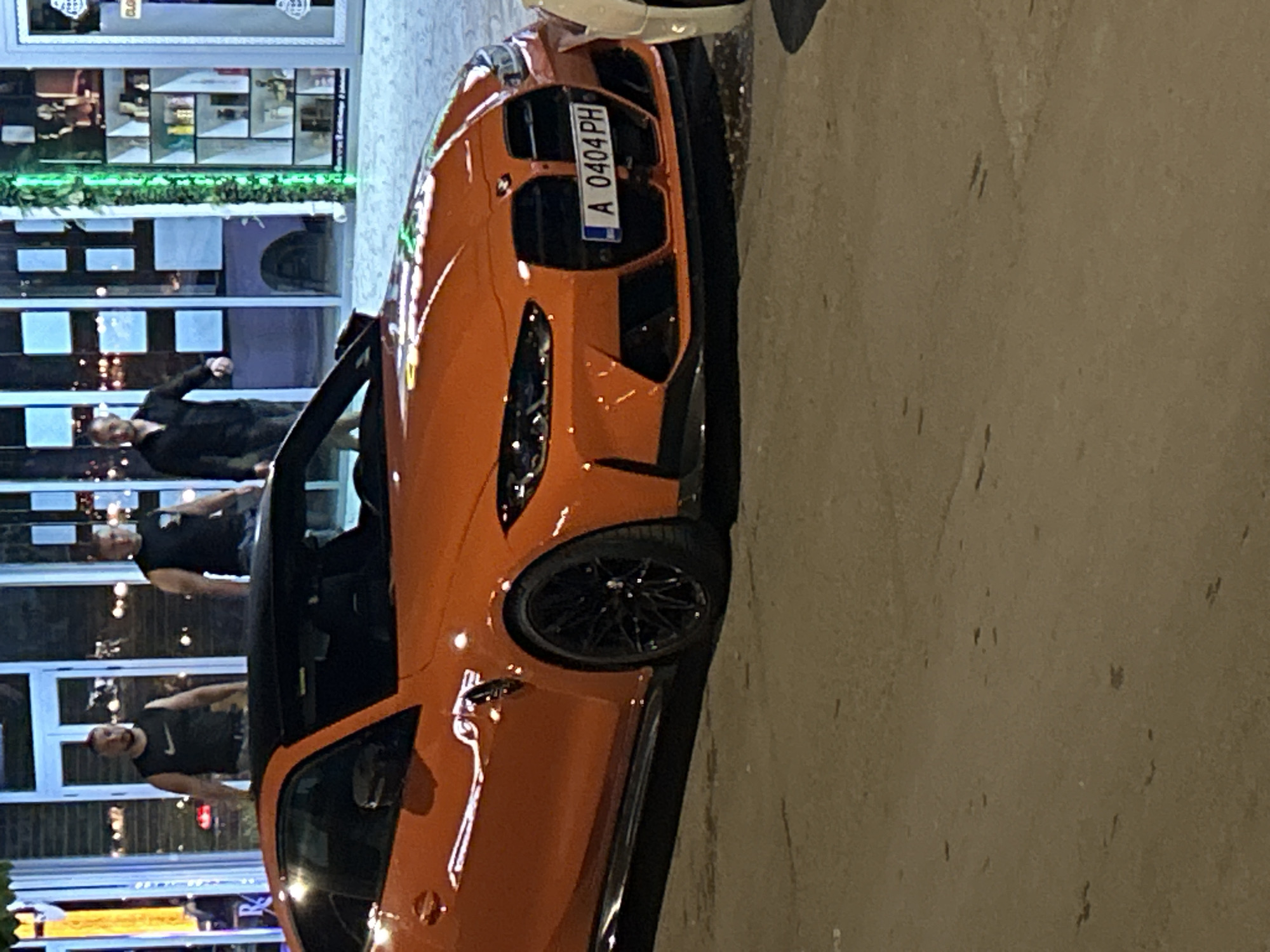 An orange sports car is parked on a street next to a building in Nesebar, Bulgaria. The car's license plate reads "A 0404 PH". Three men are reflected in the building's windows. The building has a storefront with multiple display cases and architectural details. The pavement is visible next to the car.