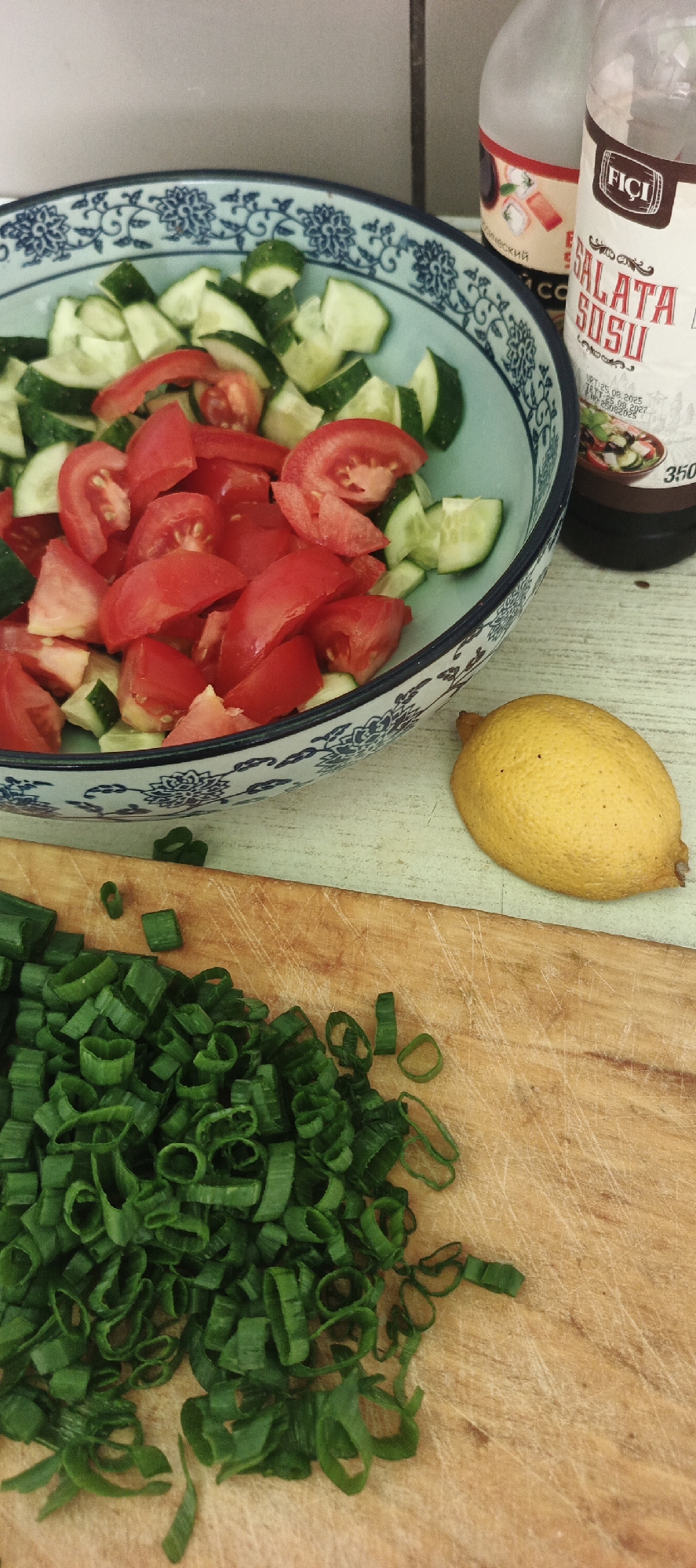 A rustic wooden table serves as the backdrop for a preparation of fresh salad ingredients. A light blue ceramic bowl, adorned with a traditional dark blue floral pattern, is filled with chopped tomatoes and sliced cucumbers. Beside the bowl, a whole, bright yellow lemon rests, its skin textured with a slightly bumpy surface. In the foreground, a rough-hewn wooden cutting board holds a generous pile of finely chopped green onions, their vibrant green contrasting with the natural wood grain. Behind the salad bowl, two bottles are visible. One is a clear glass bottle with a label featuring a prominent red and white "Fiçi" logo, indicating it's likely a food product. Another bottle, partially obscured, appears to be a dark liquid, possibly a sauce or dressing. The overall scene suggests a home kitchen setting, with the focus on fresh, raw ingredients ready to be combined. There are no people or animals present. The lighting appears to be natural, casting soft shadows and highlighting the textures of the food and surfaces. The image does not provide any specific information to determine its location in the city of Chaikovsky, Russia.