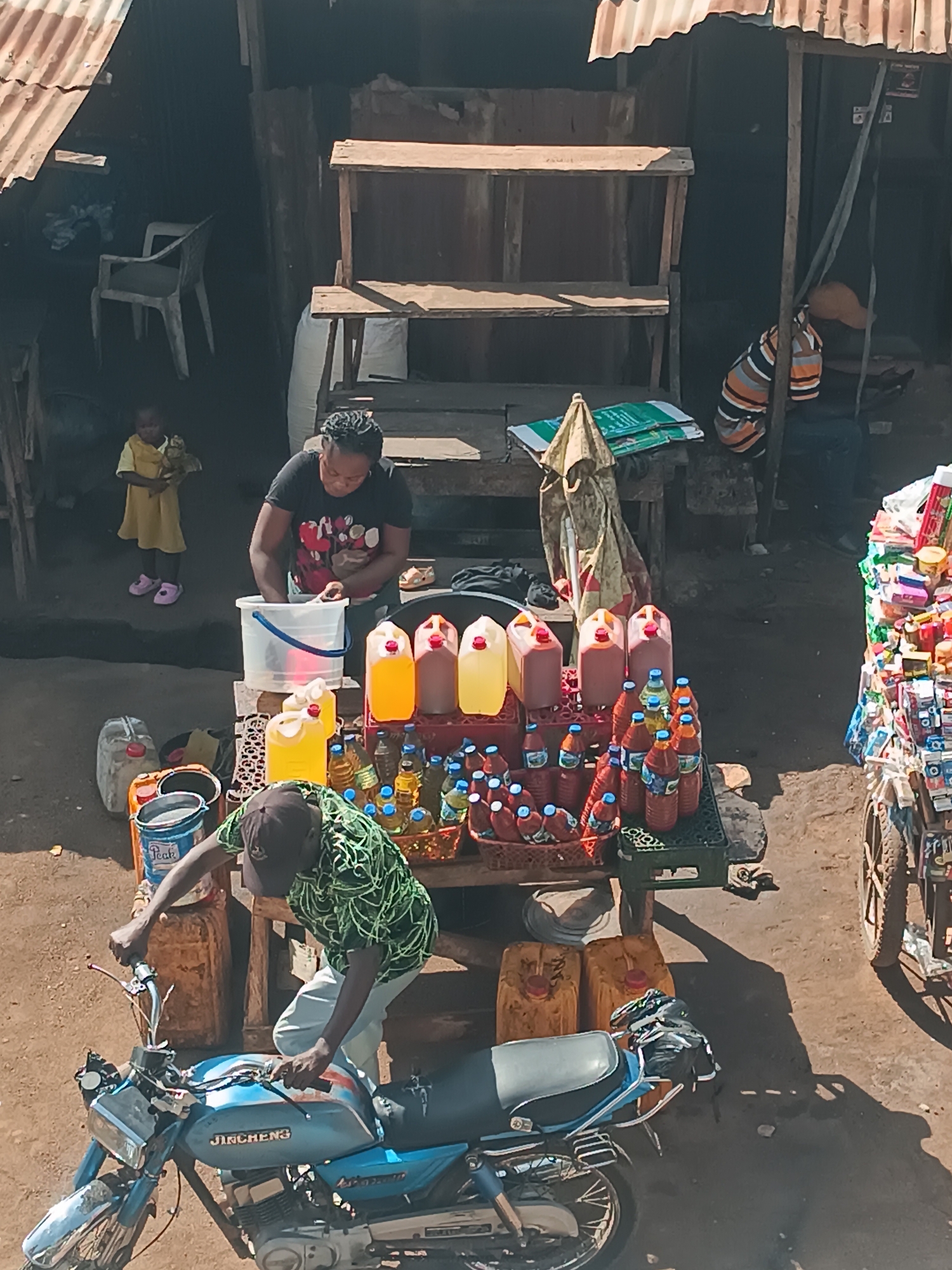 The image shows a street scene in Bukuru, Nigeria, taken from an elevated perspective. A woman stands behind a table displaying various liquids in plastic containers and bottles, possibly for sale. She is using a plastic bucket. A man in a green shirt is bending over a blue motorcycle. A young girl in a yellow dress stands near an open doorway. Another person is seated, leaning against a wooden structure to the right of the woman's stand. The environment is a mix of dirt and paved surfaces. To the right, there is a cart with a wheel that is loaded with stacks of goods.