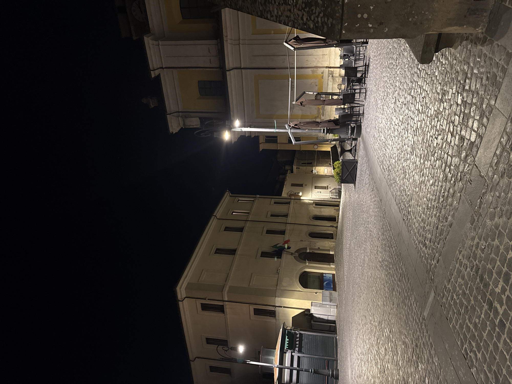 The image depicts a deserted cobblestone town square at night in Castelnuovo di Porto, Italy, illuminated by artificial streetlights. On the left, a multi-story, light-colored building with multiple windows and an arched ground-floor entrance displays an Italian flag and a regional flag. Adjacent to this building, a closed dark-green kiosk stands beneath an ornate black lamppost. On the right, a large, light-colored building, possibly a church or civic structure, features prominent yellow rectangular panels, classical architectural elements, and a visible clock face near the upper right corner. Directly in front of this right building, an outdoor seating area consists of several dark tables and chairs with three large, dark-brown, folded patio umbrellas. A dark planter containing green foliage is also present near the seating. The immediate lower right foreground is occupied by a large, textured stone block. No individuals, vehicles, or animals are present in the scene.