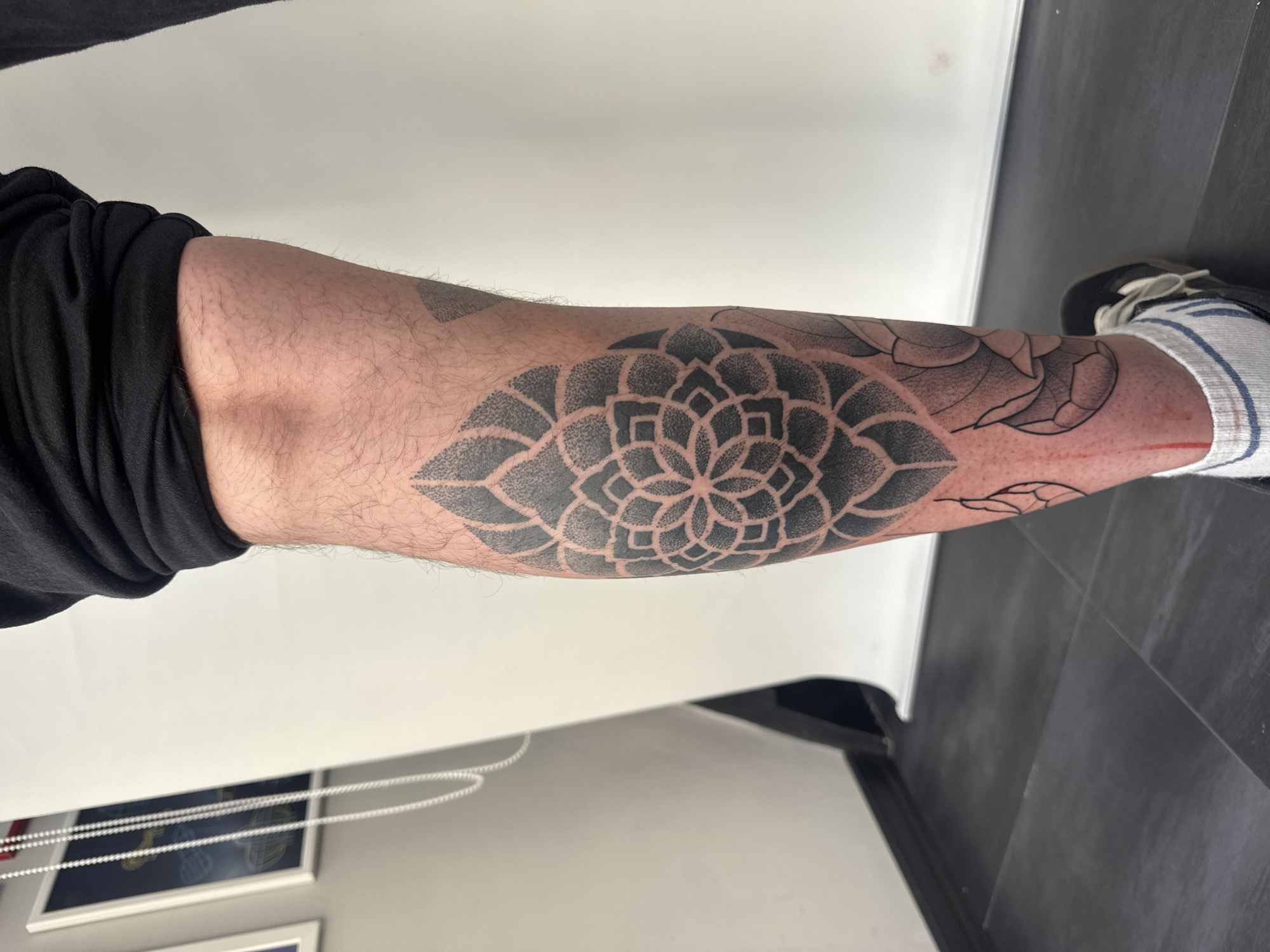 A close-up, eye-level shot shows the lower leg of a person with a freshly tattooed black mandala design on their shin. The tattoo is detailed with stippling and geometric patterns, appearing slightly red and possibly bandaged below the ankle. The person is wearing black shorts rolled up, revealing the upper part of the tattooed leg, and black sneakers with white and blue striped socks. The background is a plain white wall with some framed artwork visible on the left side. The floor is dark gray tiled. There are no visible indications of the location being in Sofia, Bulgaria.