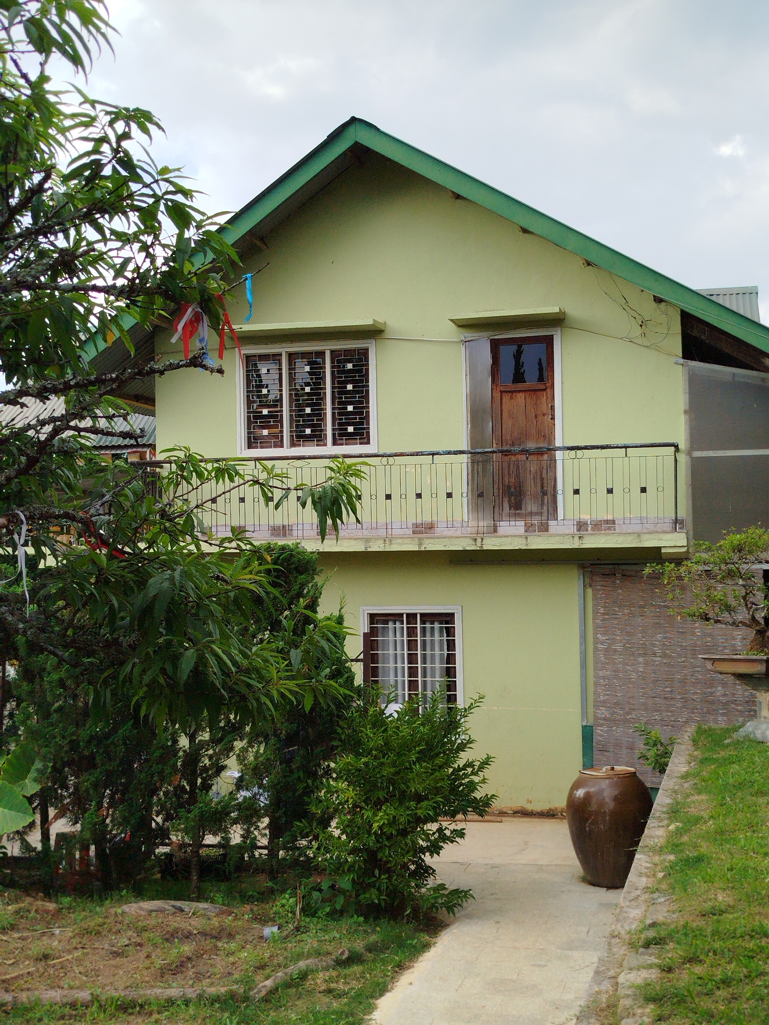 The image shows a two-story house in Da Lat, Vietnam. The house is painted a light green color with a green roof. The upper story features a small balcony with a wrought iron railing. A single wooden door is on the balcony. A window with white trim is to the side of the door. Below is another window with white trim, partially obscured by shrubbery. A large brown jar is situated on a concrete walkway adjacent to the house. The walkway is bordered by a grassy area. A tree with hanging red and blue ribbons is visible on the left side of the image. The sky is overcast.