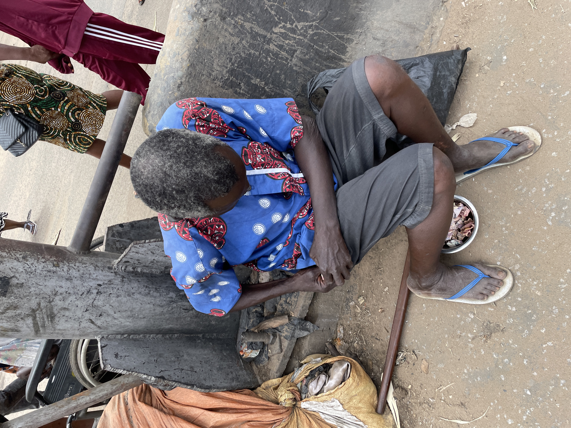 An overhead, medium shot captures a man in a blue, patterned shirt and gray shorts crouching on a dirt and gravel surface in Onitsha, Nigeria. He is positioned next to a large, dark, industrial metal structure and a sack of what appears to be fish wrapped in orange and yellow plastic. The man's gray hair is short and textured. He is leaning forward, looking down at his hands, which are clasped. His right foot is bare, wearing a light-colored flip-flop with blue straps, resting on the ground. To his right, there is a small white bowl containing what appears to be food or scraps. Behind him, a black plastic bag is visible. In the background, other individuals are present, including a woman in a colorful patterned dress and a person wearing a burgundy tracksuit with white stripes. A wheelchair is also partially visible in the background. The scene suggests a busy urban environment, possibly a market or roadside area.