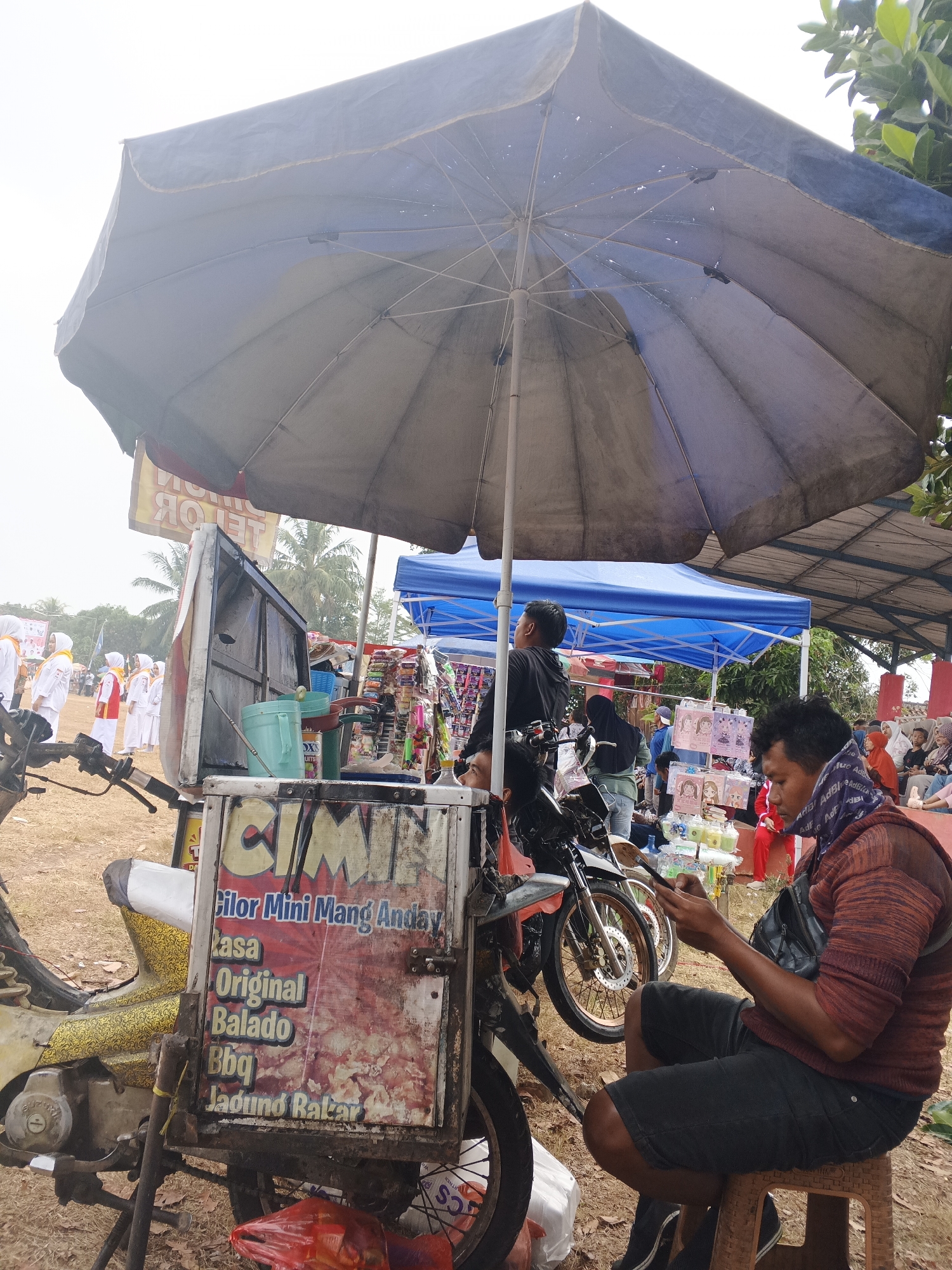 The image depicts an outdoor scene in the city of Warungkiara, Indonesia, likely during a local event or market day. The foreground features a food cart attached to a motorcycle, with a large, dark umbrella providing shade. The cart displays a menu with handwritten text in Indonesian, listing food items such as "Cilor Mini Mang Anday," "Rasa Original," "Balado," "Bbq," and "Jadund Bakar." A man, wearing a patterned scarf and a maroon hooded sweatshirt, sits on a stool to the right of the cart, looking down at his phone. In the background, several stalls are visible, including one with a blue canopy displaying various packaged goods, possibly snacks or small toys. Further in the background, a group of people, some wearing white and orange attire, are gathered, suggesting a procession or a performance. The ground is covered with dry grass and dirt, and palm trees are visible in the distance, indicative of the tropical climate. The overall atmosphere appears to be that of a bustling community gathering.