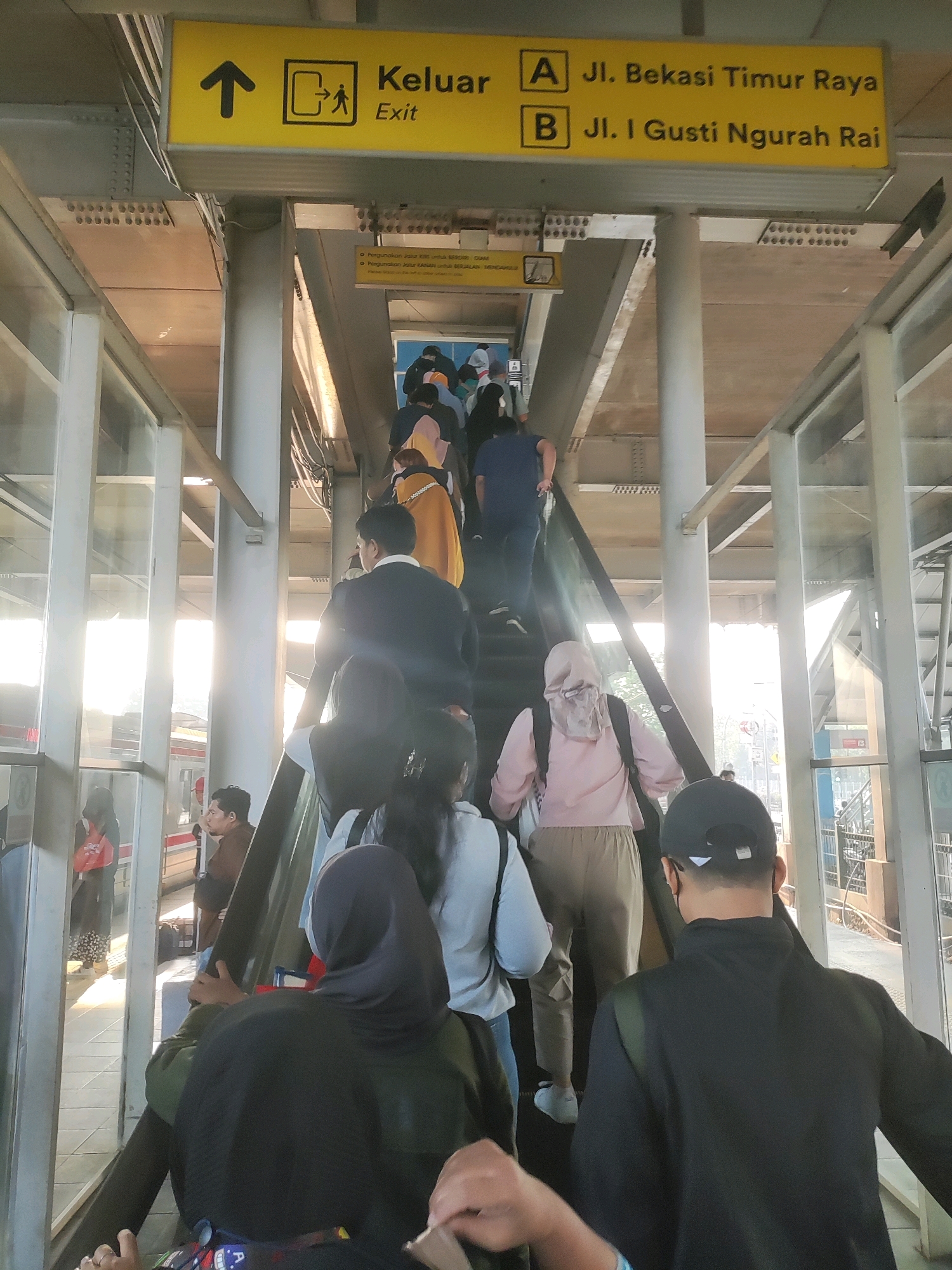 An upward-facing, eye-level shot captures a bustling escalator within a transit station in East Jakarta, Indonesia. The escalator, flanked by glass railings and industrial-style concrete pillars, ascends towards a bright, sunlit exit. A prominent yellow directional sign, positioned above and to the left of the escalator, indicates "Keluat Exit" with an arrow pointing upwards, and further displays two routes: "A Jl. Bekasi Timur Raya" and "B Jl. I Gusti Ngurah Rai."
A diverse group of individuals, predominantly dressed in casual attire, are ascending the escalator. Many are wearing headscarves, suggesting a conservative cultural context. The sun, shining brightly from the left side of the frame, casts long shadows and creates a lens flare effect, illuminating the scene with a warm glow. The background reveals elements of the station's architecture, including elevated tracks and what appears to be a train, hinting at the station's function as a public transportation hub. The overall atmosphere is one of movement and daily transit, characteristic of a busy urban environment.
