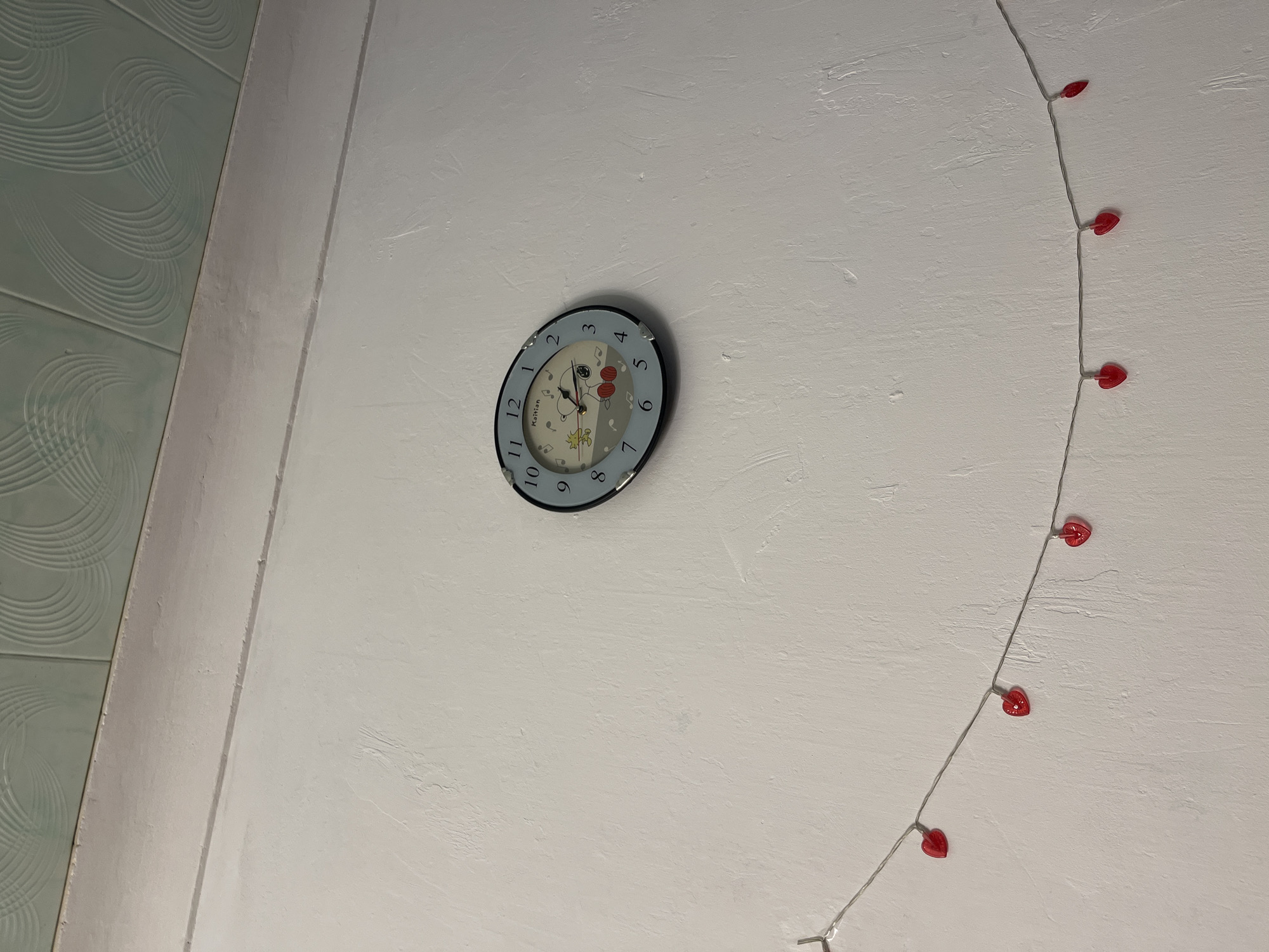 A close-up interior shot shows a light gray wall. A child's clock with a blue rim and cartoon characters is affixed to the wall. Adjacent to the clock is a string of lights shaped like red hearts. A light blue wall with a textured, swirling pattern is visible on the left edge of the frame. The location is identified as Perm, Russia.