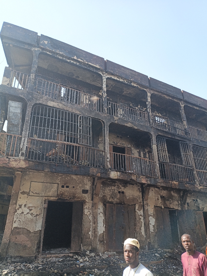 The scene in Fagge C, Nigeria, depicts a three-story building severely damaged by fire. The entire facade is heavily charred, exhibiting extensive blackening, peeling paint, and plaster that exposes the underlying masonry. Each of the three visible levels features a continuous balcony with remnants of burnt railings and exposed, empty window and door apertures. Debris, consisting of ash and rubble, covers the ground immediately in front of the structure. Two adult males are positioned in the foreground. The individual on the left wears a light-colored shirt and a yellow skullcap, looking directly forward. The individual on the right wears a maroon top and is positioned slightly behind and to the right of the first, also facing forward. Both individuals appear to be observing the damaged environment under a clear sky.