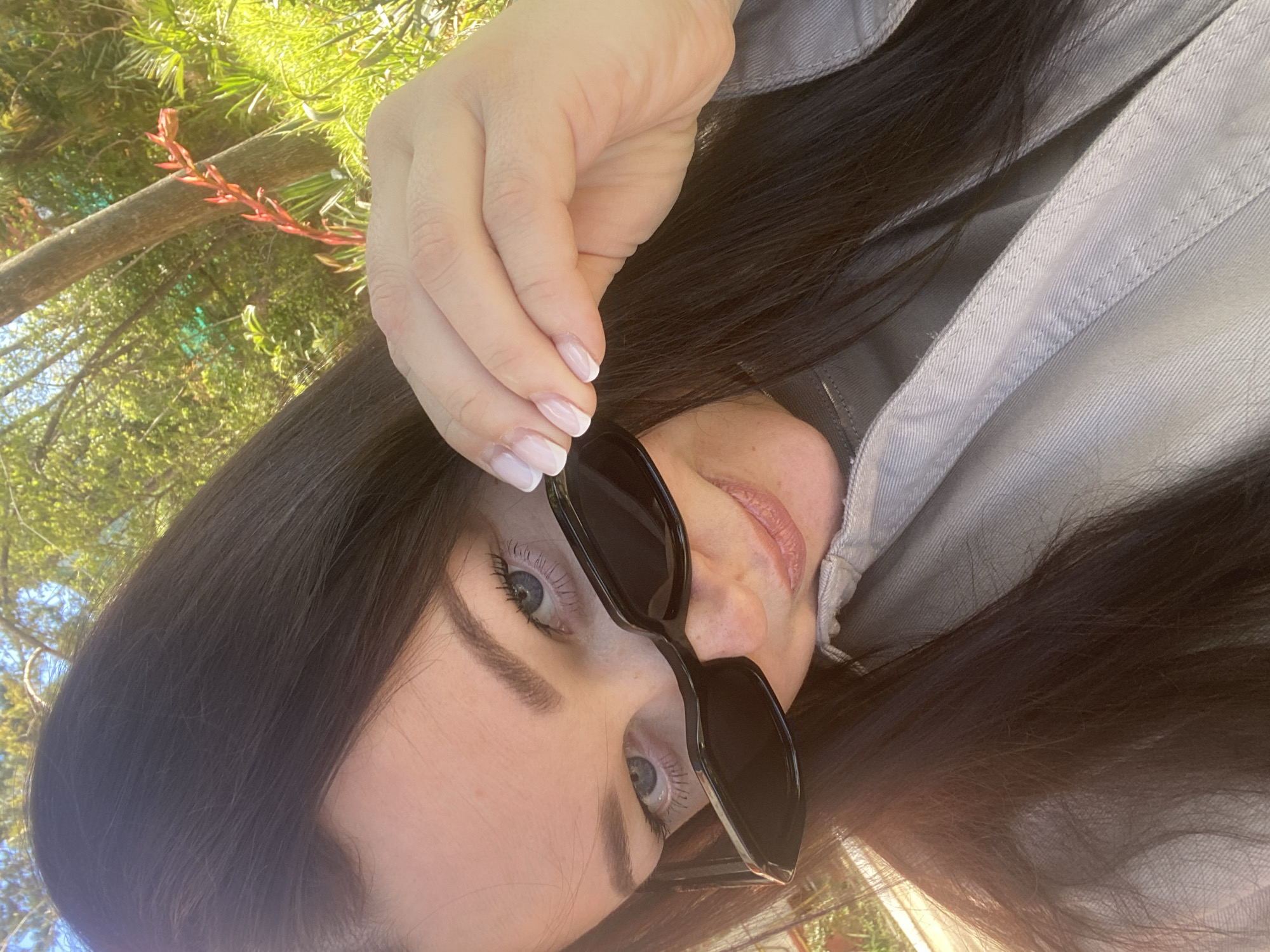 A close-up, high-angle outdoor portrait features a woman, likely taken in Sochi, Russia, on a bright, sunny day. The image is rotated, showing the woman's head tilted to the right. She has long, dark hair that cascades around her face and over the shoulder of a light grey jacket, which appears to be denim or a similar material with visible stitching.
The woman is holding a pair of black, rectangular sunglasses just below her eyes, revealing her striking light blue eyes, dark eyelashes, and well-groomed eyebrows. Her lips are slightly parted, showing a natural or light pink hue. Her right hand is visible in the upper left, positioned near her forehead, showcasing neatly manicured fingernails with a classic French tip design (pinkish base with white tips).
The background is a vibrant blur of green foliage, indicating a lush, natural, possibly treed environment. A prominent tree branch with small reddish sprouts runs across the upper left part of the frame. Bright sunlight illuminates the scene, casting highlights on her hair and the surrounding greenery, signifying clear, sunny weather. The overall impression is a relaxed and natural moment. No text is visible in the image.
