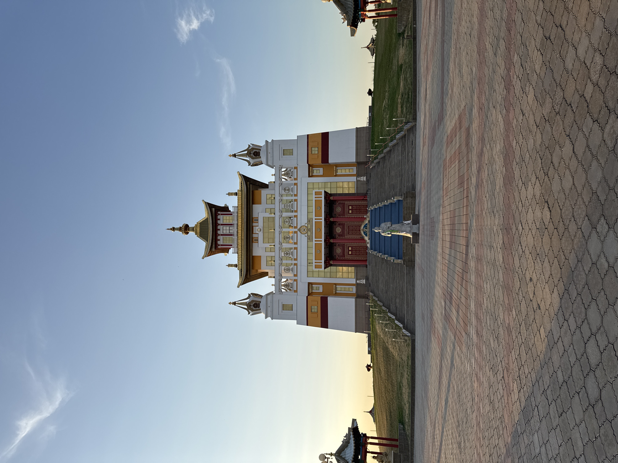 The image shows the Golden Temple of Elista, Russia, located in the Republic of Kalmykia. The temple is a large, multi-tiered Buddhist structure with a white base, yellow and red accents, and a golden roof. It is set on a grassy hill, with a wide, paved plaza in front. A statue of a serene figure stands on the plaza, facing the temple. The sky is a clear blue with a few wispy clouds, and the sun is setting, casting long shadows across the plaza. The scene is tranquil and majestic, conveying a sense of peace and spiritual contemplation.