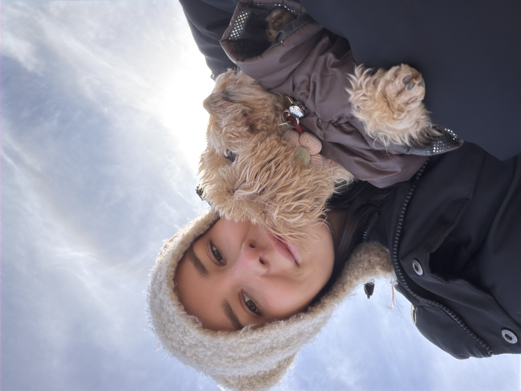 The image captures a low-angle, upward-looking perspective of a young person and a small dog against a bright, cloudy sky in Мокшино, Russia. The primary subject is a young person, appearing to be female, with light skin and dark hair. She looks directly at the camera with a soft, gentle smile. She is wearing a warm, light-beige, possibly knitted or fleece-lined, hat that frames her face and a dark winter jacket, likely black or dark blue, with a visible zipper and metal snaps.
Cradled near her shoulder and neck is a small, fluffy dog with light brown fur, possibly a terrier breed. The dog is also dressed in a warm, puffy brown winter coat or harness, and a small, shiny bell or charm is visible on its collar. Its head is slightly turned away, and its fur appears shaggy. One of the dog's paws, showing distinct paw pads, is visible near the person's shoulder.
The background is dominated by a bright, hazy sky with soft, cirrus-like clouds. The sun is intensely bright, positioned high in the frame, creating a strong backlight that illuminates the edges of the subjects. The winter clothing of both subjects suggests a cold outdoor environment during the daytime. No text is visible in the image. The scene conveys a warm, affectionate moment shared between the person and their pet.