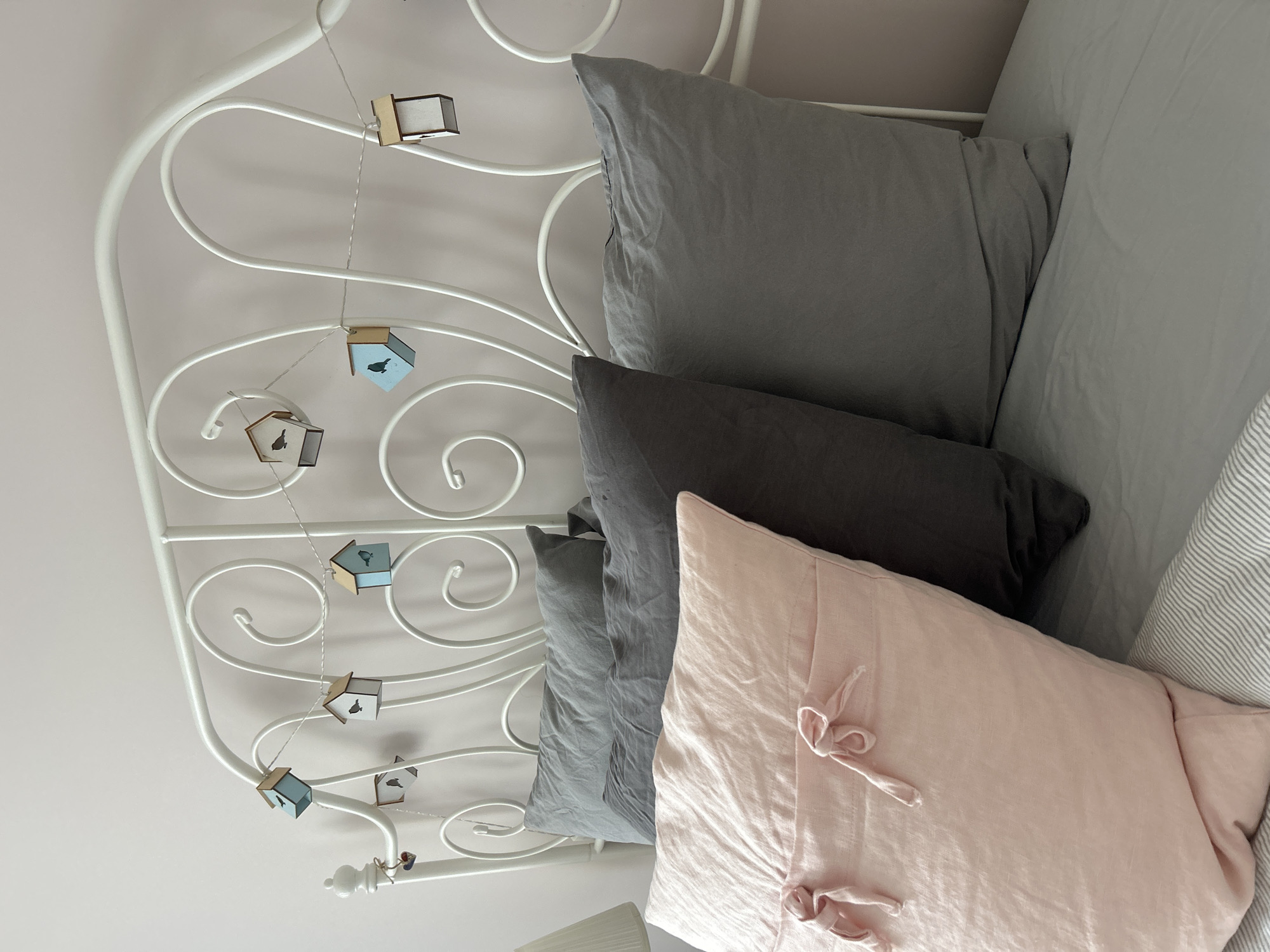A close-up, high-angle shot shows a bed with a white, ornate metal headboard. A string of decorative birdhouses is strung across the headboard. On the bed are several pillows: a light gray pillow at the top, followed by a dark gray pillow, and a pink pillow at the bottom with ties. The bed linens have a subtle stripe pattern. The scene is likely an interior setting; no specific location within Plovdiv, Bulgaria, can be determined from the image. There are no people visible in the image.