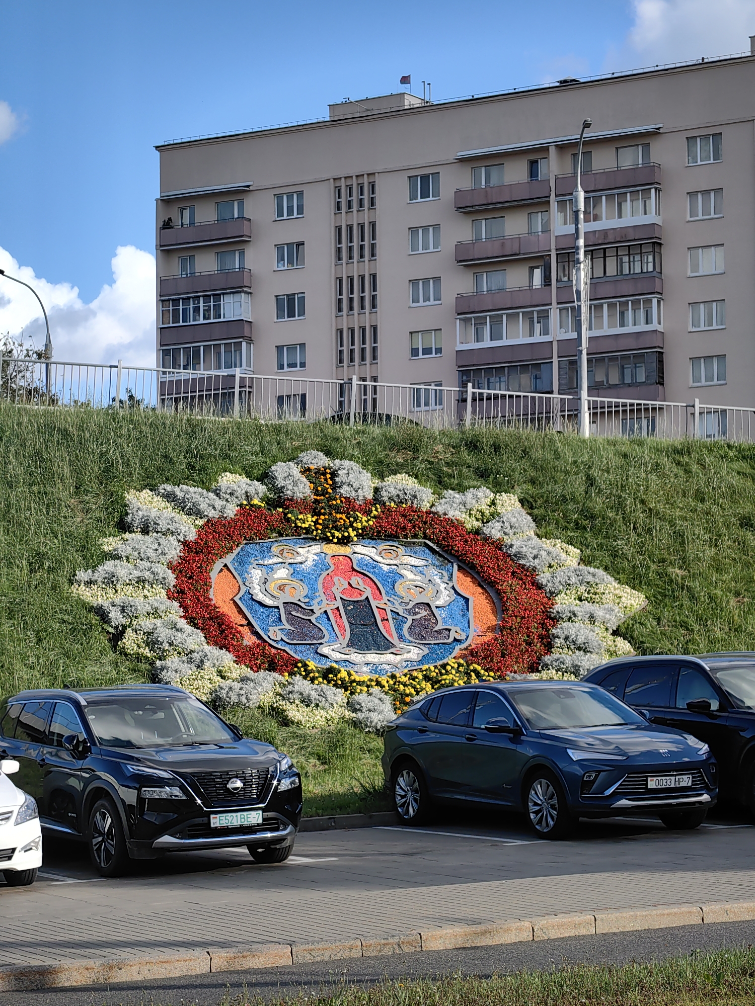 The image depicts a large floral arrangement in the shape of a mosaic on a grassy embankment, located in Minsk, Belarus. The mosaic features a blue background with a central image of two figures, one appearing to be holding a child or an object. Surrounding the mosaic is a colorful border of red, orange, yellow, and white flowers, with green grass covering the embankment. In the foreground, a black Nissan X-Trail SUV and a dark blue Buick Encore are parked in a lot paved with concrete slabs. The license plate of the Nissan is visible as E521BF-7, and the license plate of the Buick is 0033 HP-7. Behind the embankment, a multi-story residential building with numerous windows and balconies is visible under a bright blue sky with scattered white clouds. A street lamp and a white metal fence are also present.
