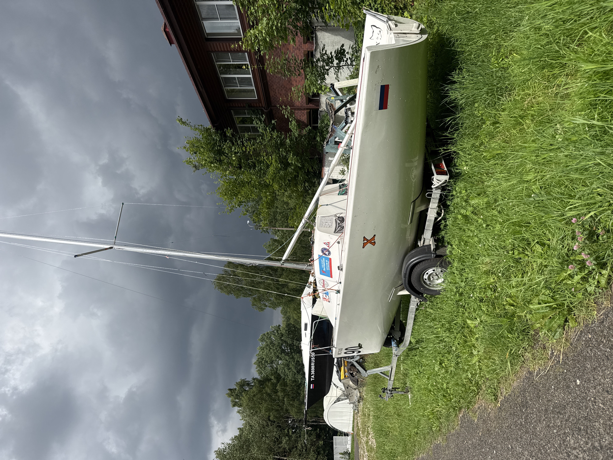 In Povedniki, Russia, on a cloudy day, a white sailboat rests on a trailer on a grassy area next to a paved path. The sailboat has a sail mast and various markings and stickers on its hull, including the number "90" and a Russian flag. In the background, a dark-colored sailboat is visible behind a white tent-like structure. Further back, a two-story brick building with windows stands amongst lush green trees. The sky is filled with dark, dramatic clouds.