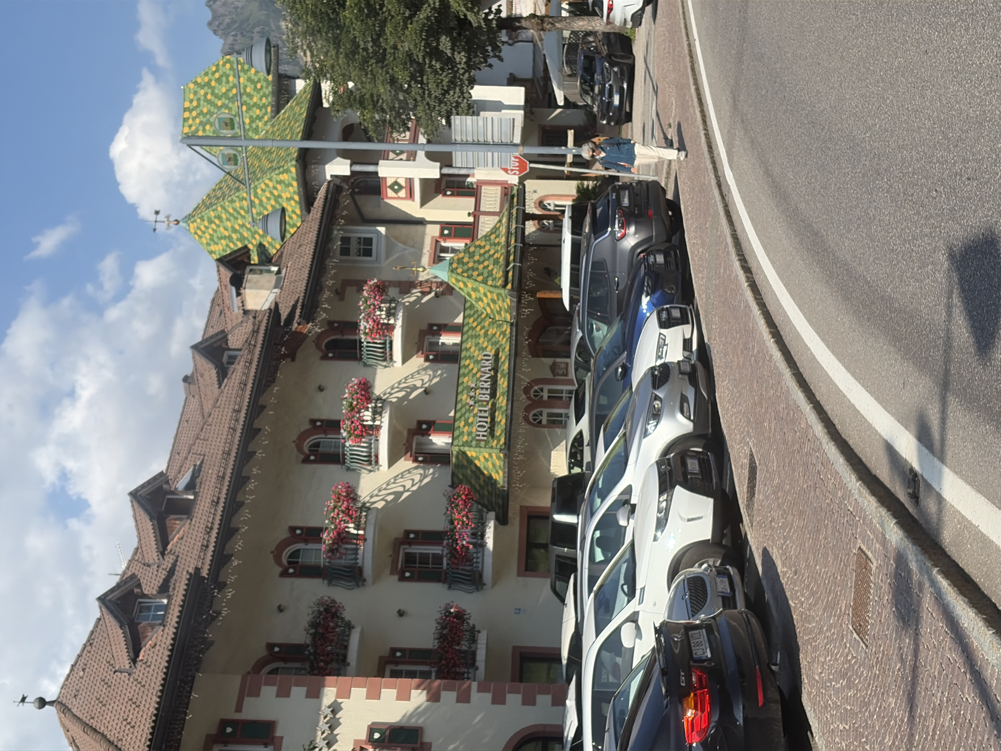 The image shows a street scene in Canazei, Italy. In the foreground, several cars are parked along a cobblestone sidewalk next to a paved road. A woman is walking on the sidewalk, facing away from the camera. Behind the cars, a multi-story building with a distinctive green and yellow tiled roof stands prominently. The building's facade is cream-colored with red accents around the windows and balconies adorned with blooming red flowers. A sign above the entrance reads "HOTEL BERNARD". The background features a clear blue sky with scattered clouds and a glimpse of distant mountains. The overall impression is a picturesque street in a European town, likely in the Alps, given the architecture and mountainous backdrop.