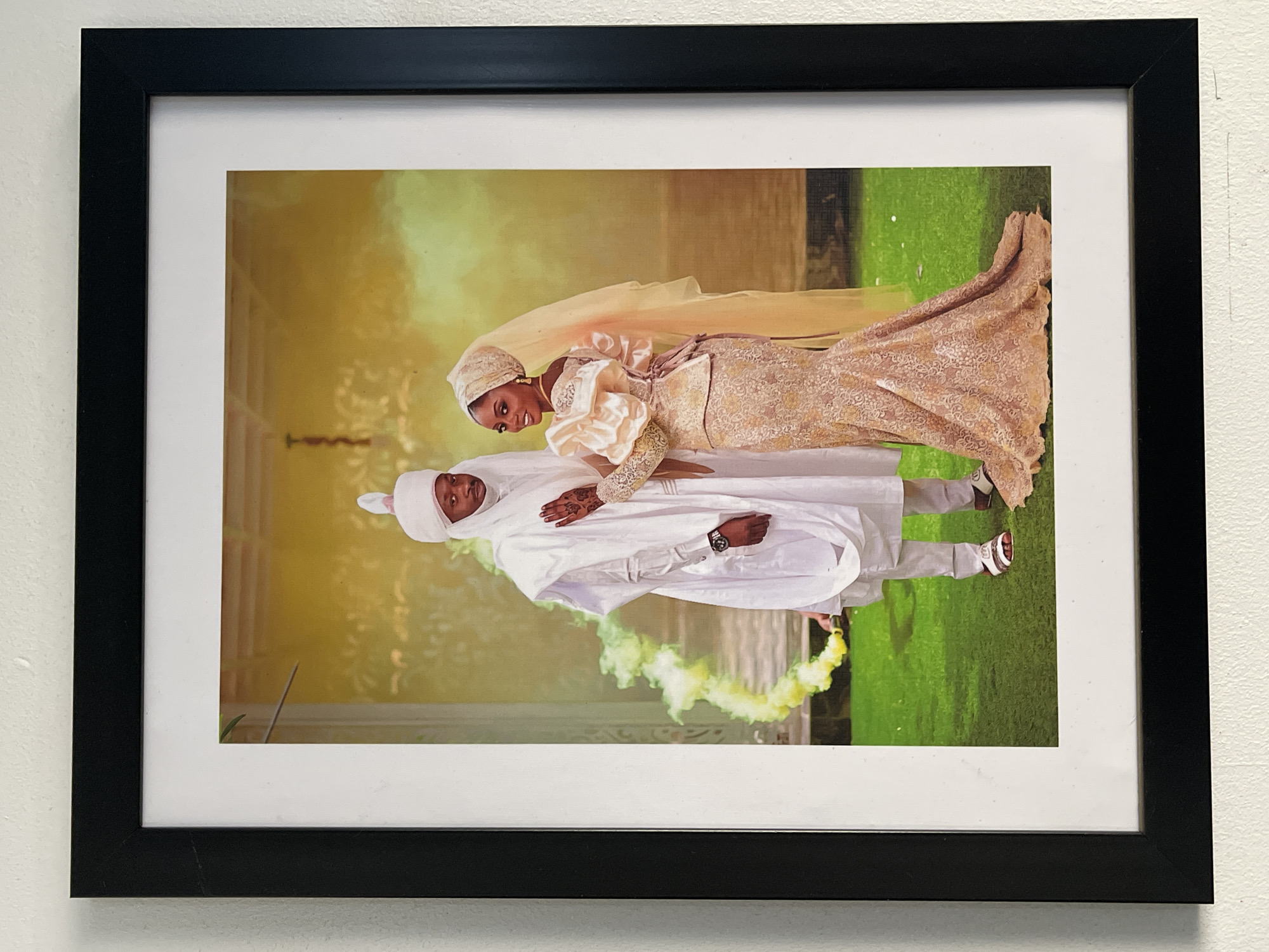 This media file features a framed photograph of a man and a woman, likely a couple, posing for a picture in an outdoor setting. The black frame encloses a white mat, which in turn frames the photograph.
The subjects are dressed in formal, traditional attire. The woman, positioned on the right, wears an elaborate light peach or gold-colored gown with intricate lace patterns, a matching headscarf, and a long veil. She has visible earrings and elaborate henna designs on her left hand. Her expression is joyful, with a bright smile. The man, on the left, is clad in a white traditional outfit, possibly an Agbada, with a matching head-covering and sandals. He has a calm expression, looking forward. Both are wearing watches.
The couple stands close together, with the woman's right arm gently resting on the man's left arm. The background features a vibrant green ground, possibly artificial turf or well-maintained grass, and a lighter, yellowish-golden upper portion, which appears to be a light effect or smoke. A burst of vivid yellow smoke is visible near the man's feet, adding a dynamic and celebratory element to the scene.
The activity taking place is a formal photoshoot, indicative of a wedding, engagement, or significant celebration. The attire, henna, and overall presentation suggest a cultural context consistent with Abuja, Nigeria, as provided. There is no visible text in the image. The lighting suggests it is daytime, possibly late afternoon.