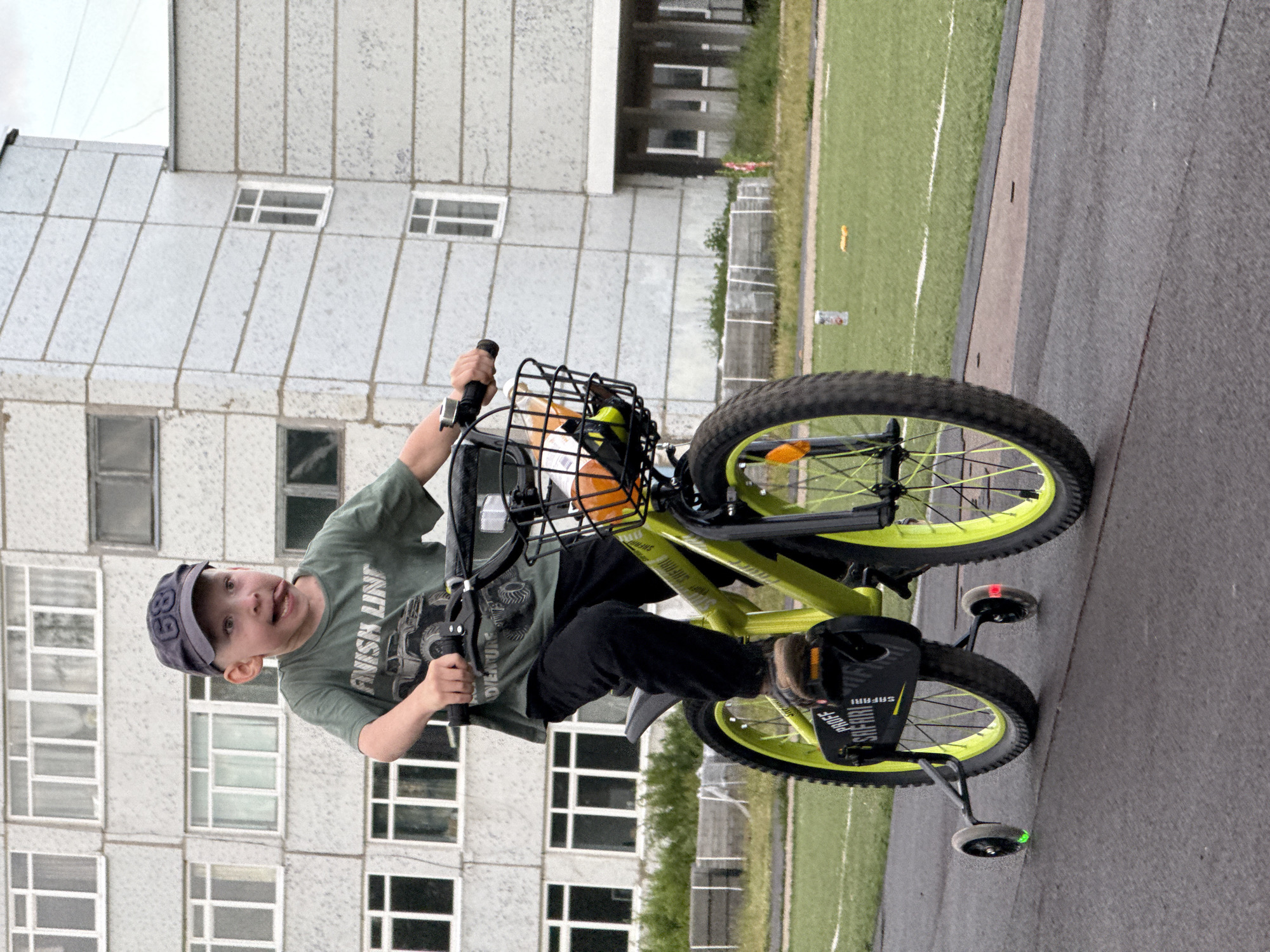 A young boy, wearing a green t-shirt and black pants, is riding a yellow bicycle with training wheels on a paved surface. He has a blue baseball cap on and is sticking his tongue out playfully. The bicycle has a black basket on the handlebars, containing an orange object and other items. The training wheels have red and green lights. In the background, there is a tall, gray, multi-story building with numerous windows. To the right of the building is a green field, possibly a sports field with white lines. The sky appears overcast. The image is oriented vertically.
Visible text includes "FINISH LINE" and what appears to be "SAFARI" and "RUOFF" on the bicycle pedals. The number "89" is visible on the boy's cap.